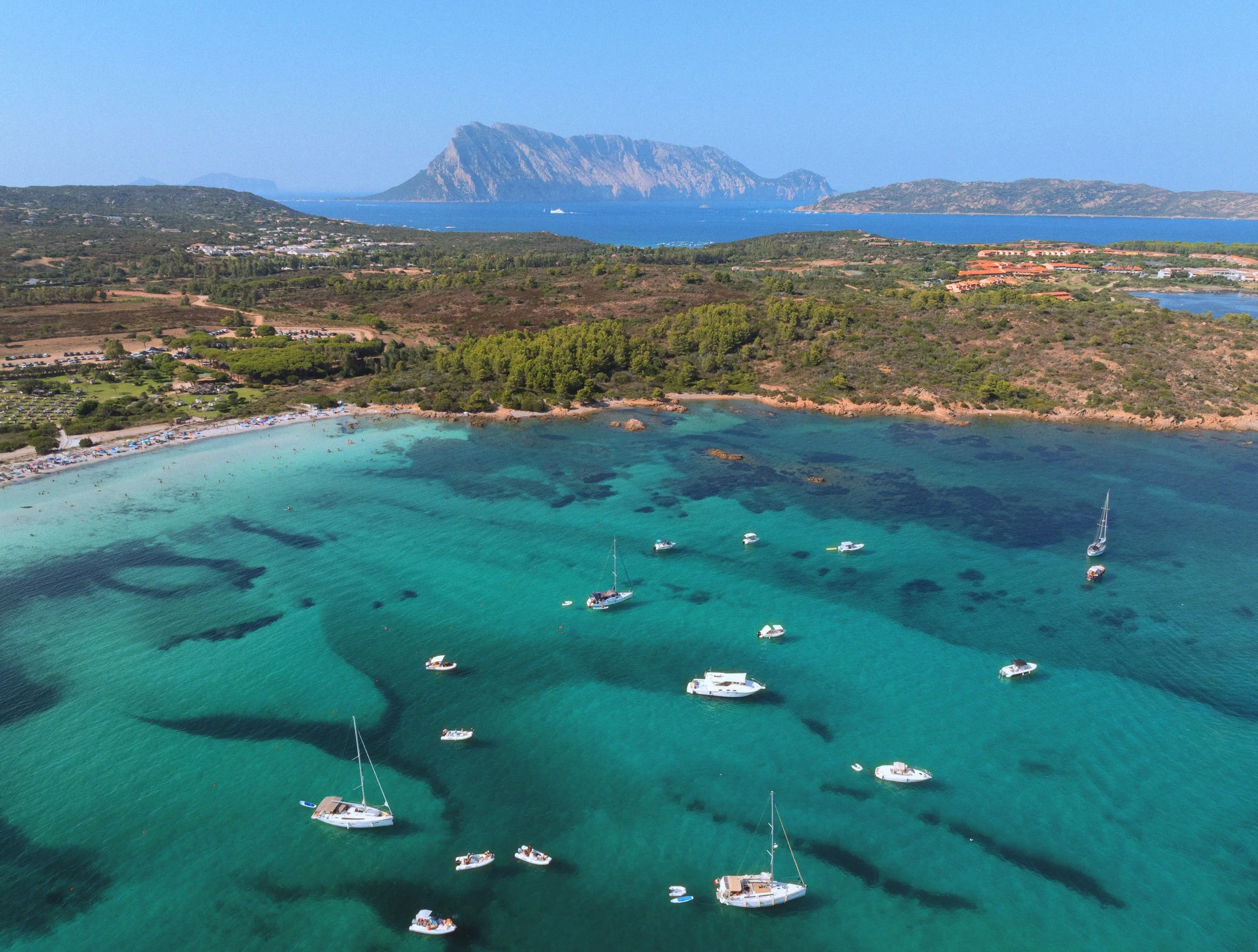 Sailboats and dinghies anchored over turquoise shallows, with Tavolara on the horizon