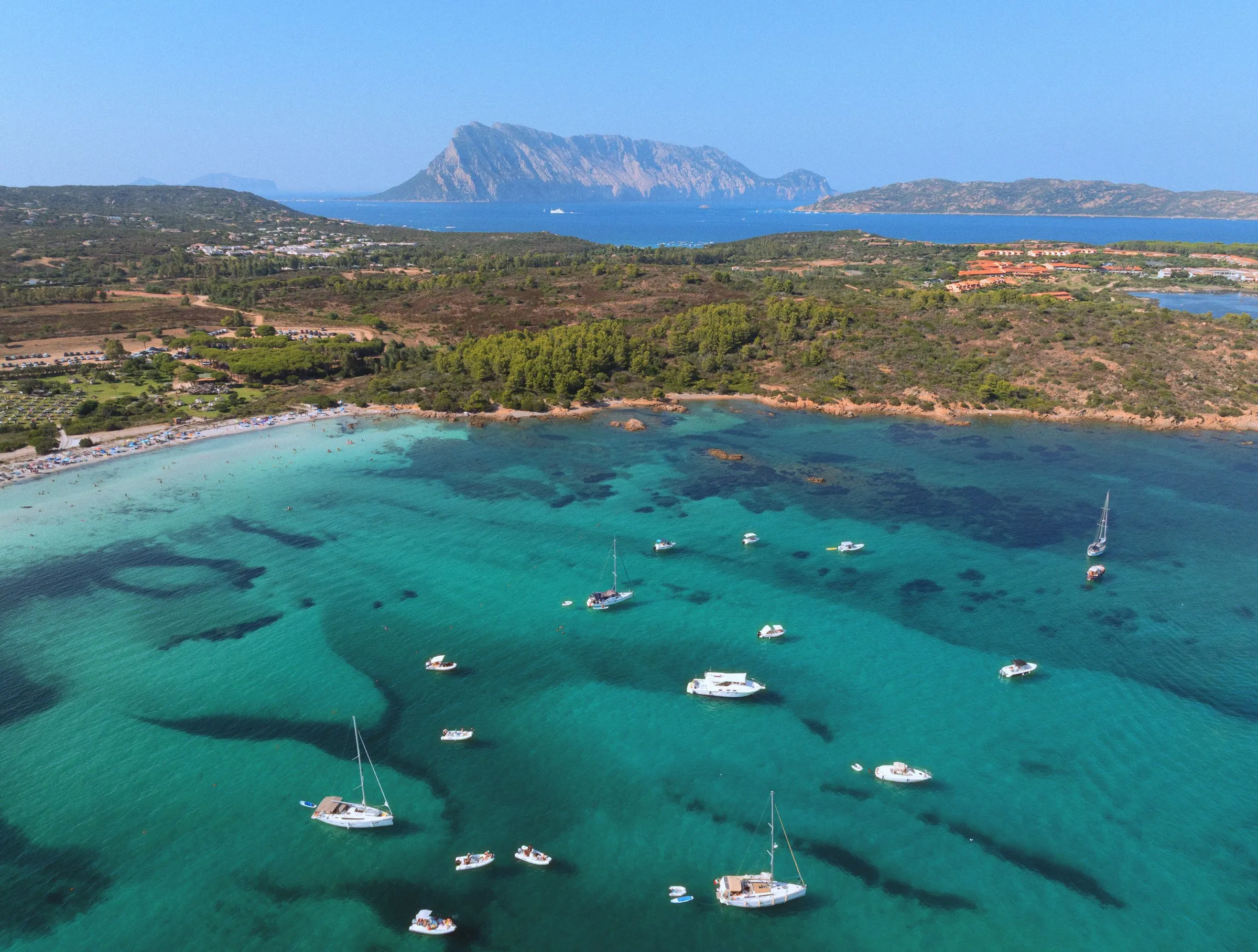 Sailboats and dinghies anchored over turquoise shallows, with Tavolara on the horizon
