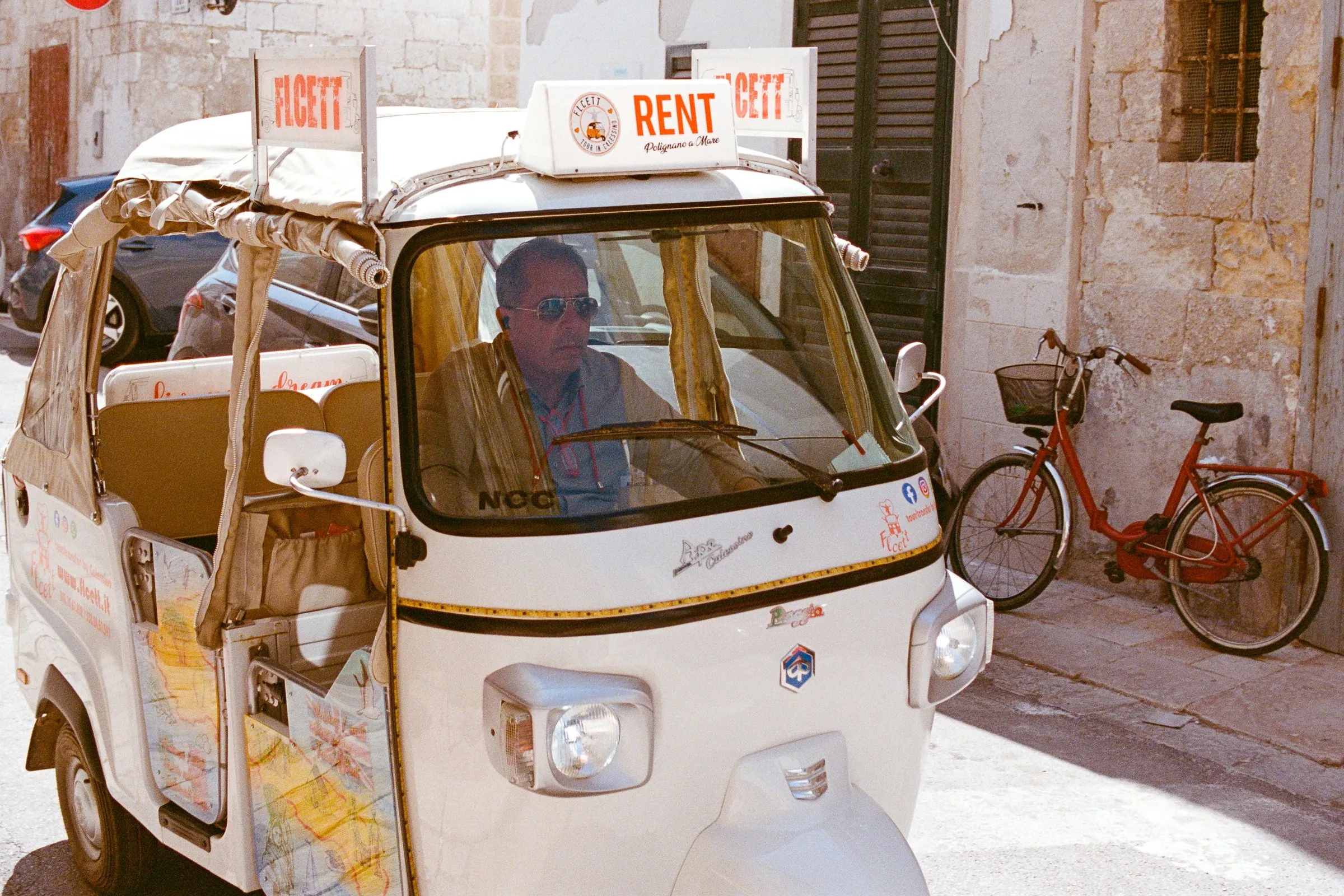 A driver waits in a white Ape tuk-tuk with a rental sign, a red bicycle parked behind