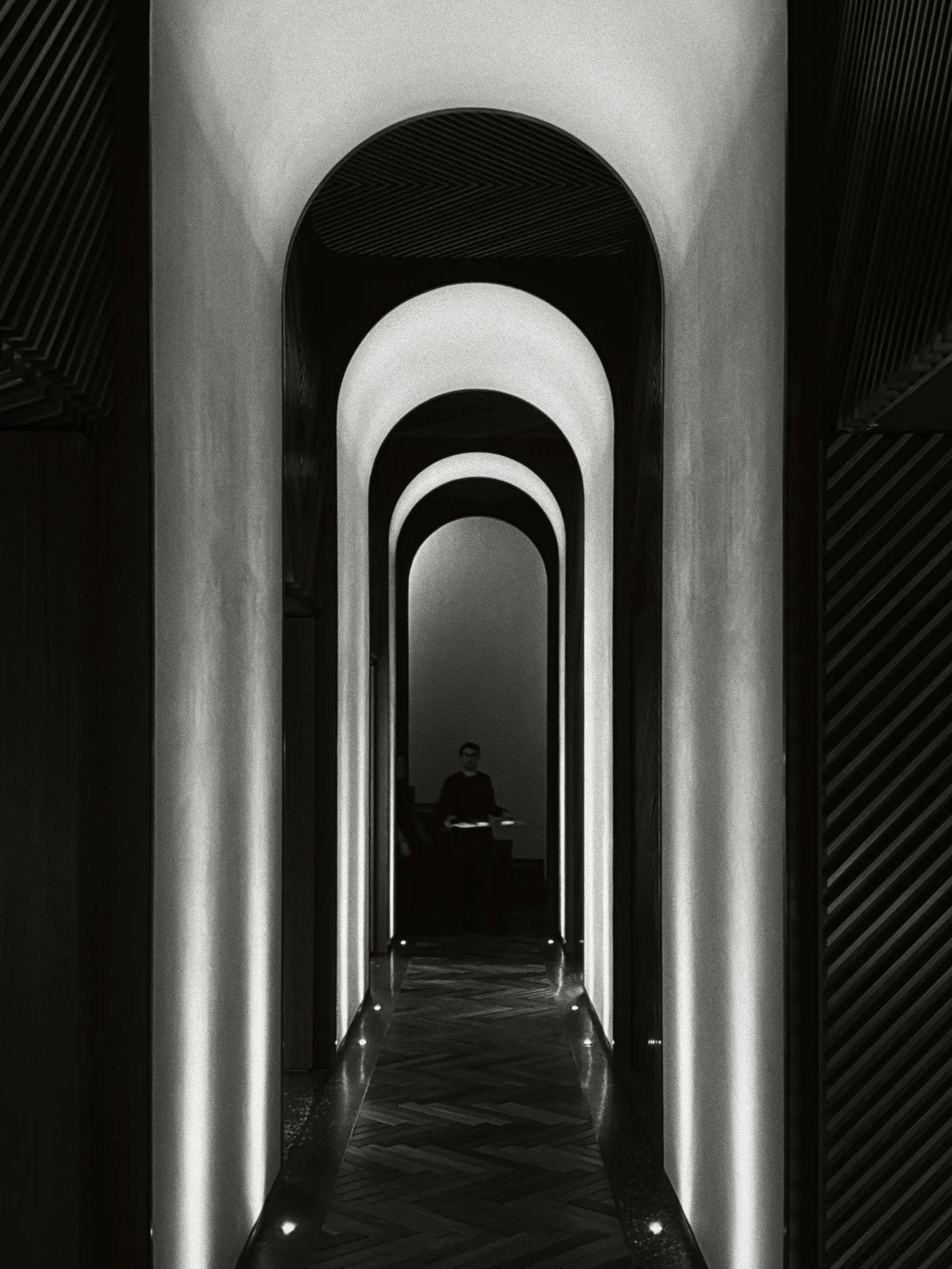 Black and white view down a corridor of lit arches to a lone waiter at the far end