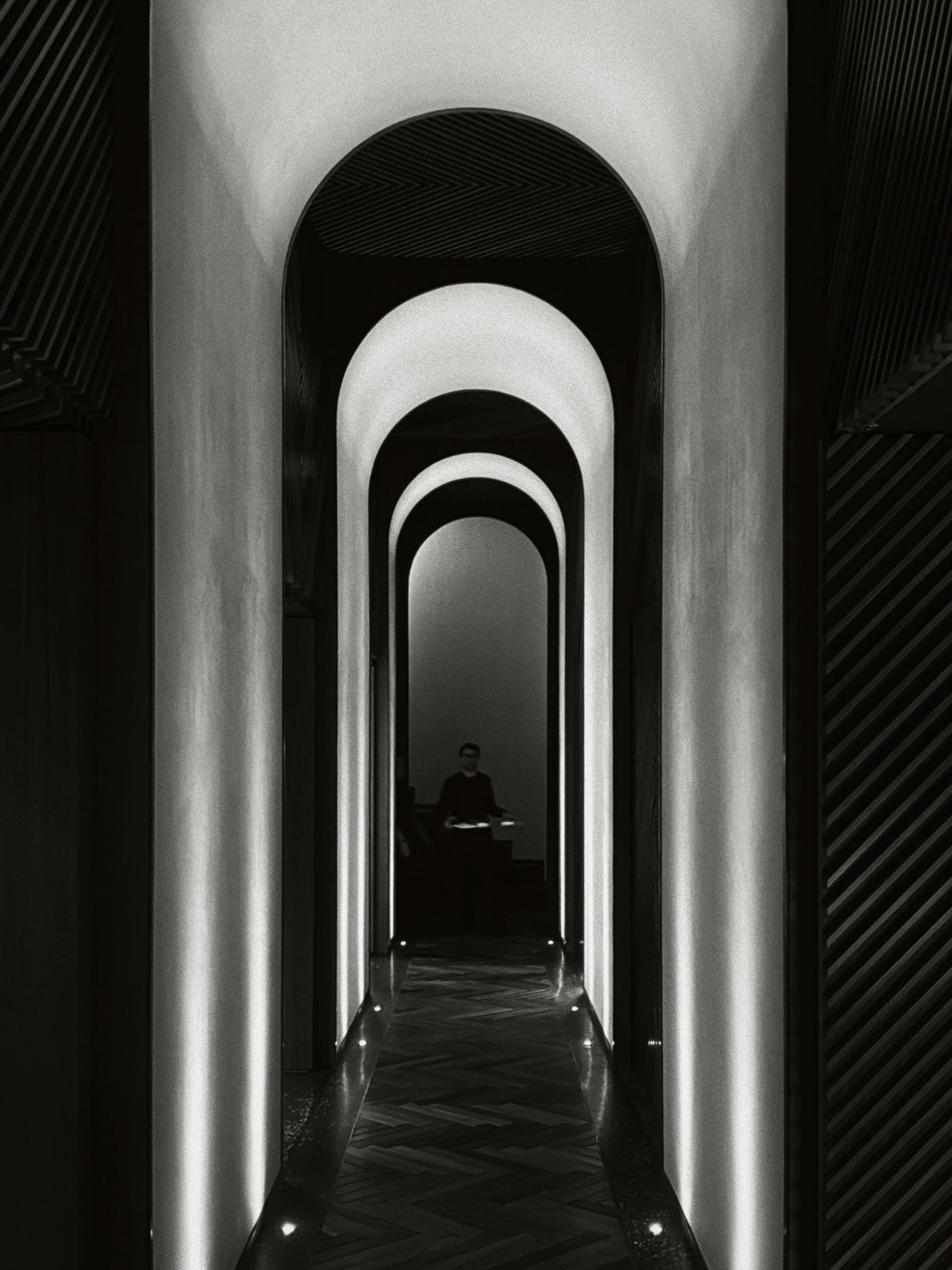 Black and white view down a corridor of lit arches to a lone waiter at the far end