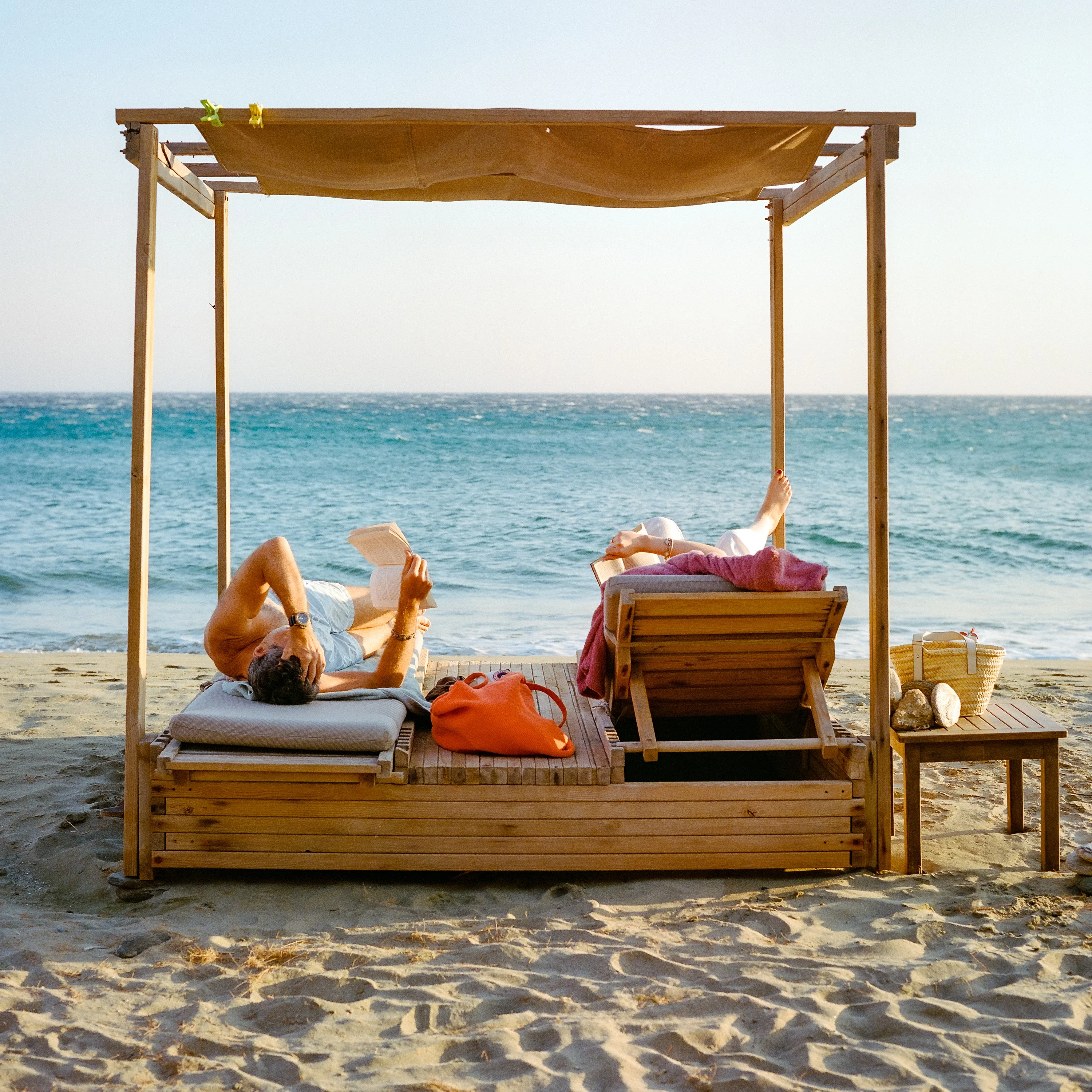 Two people reading on a canopied daybed on the sand, the sea behind
