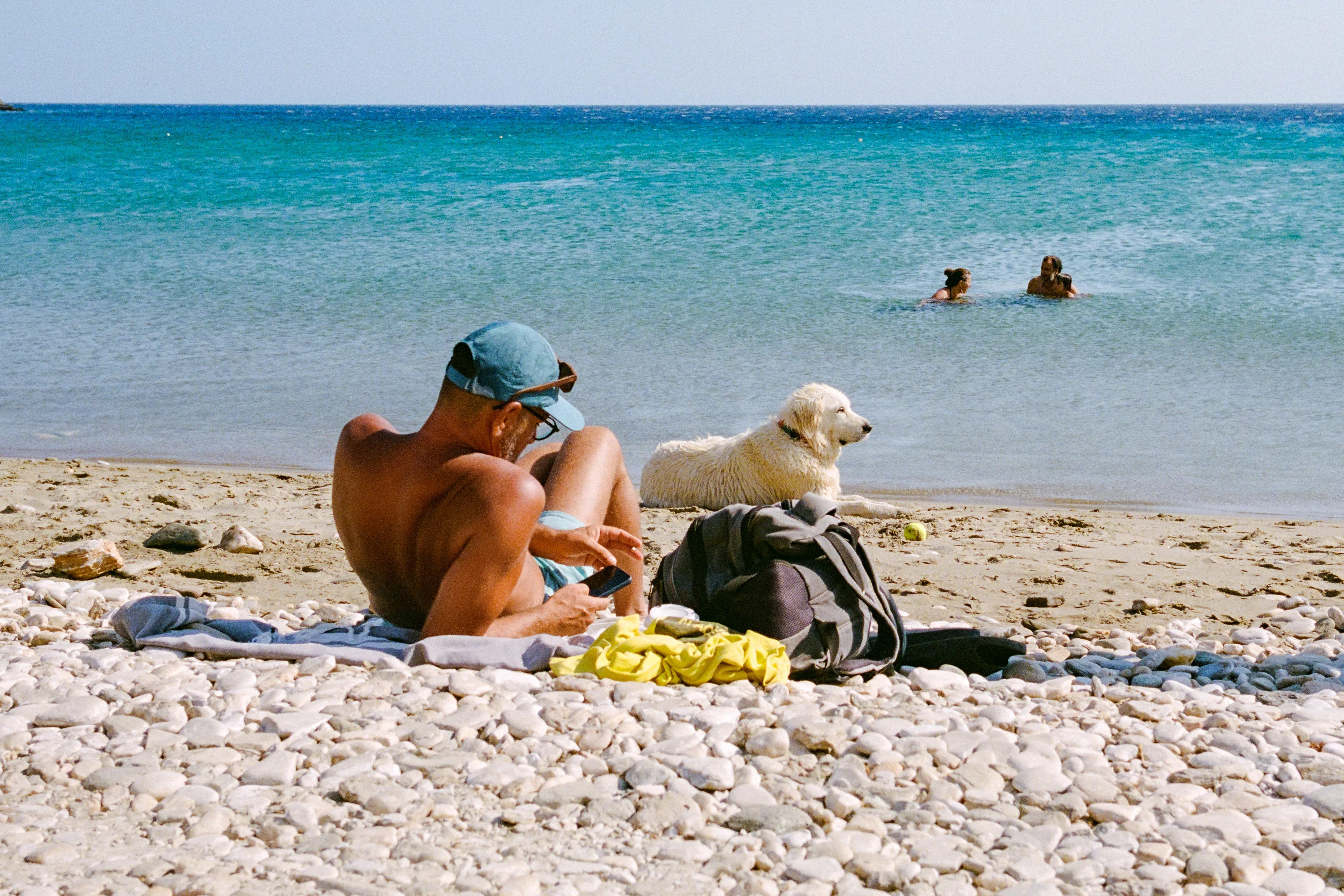 A man in a cap sitting on a pebble beach beside a white dog, swimmers out in the clear sea