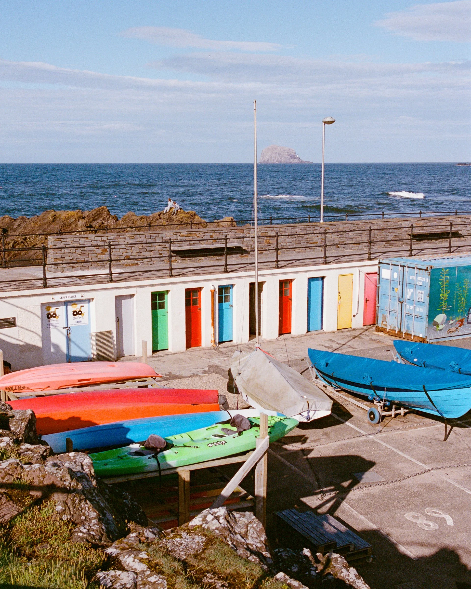 A row of brightly painted hut doors by the sea, with kayaks and boats on the rocks