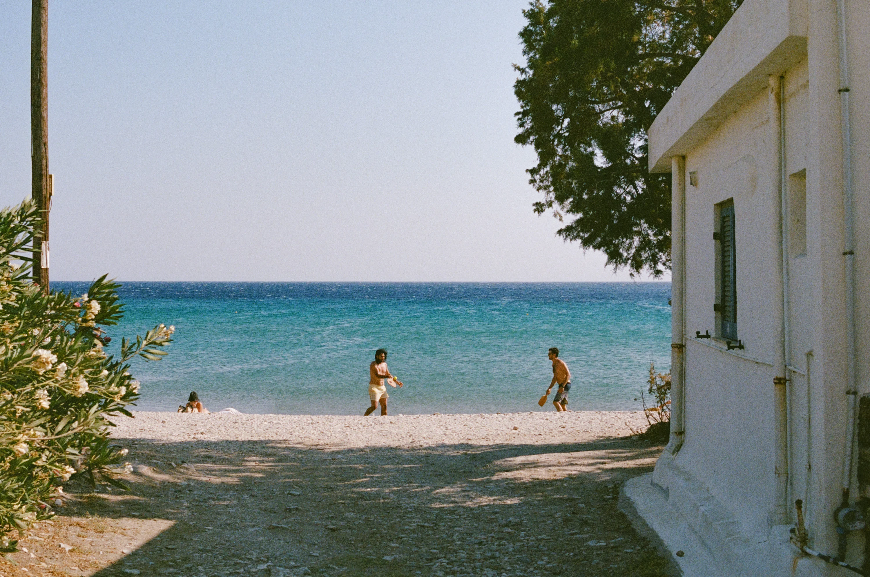 A sandy lane between a white wall and trees opening onto a turquoise sea, two people walking to the water