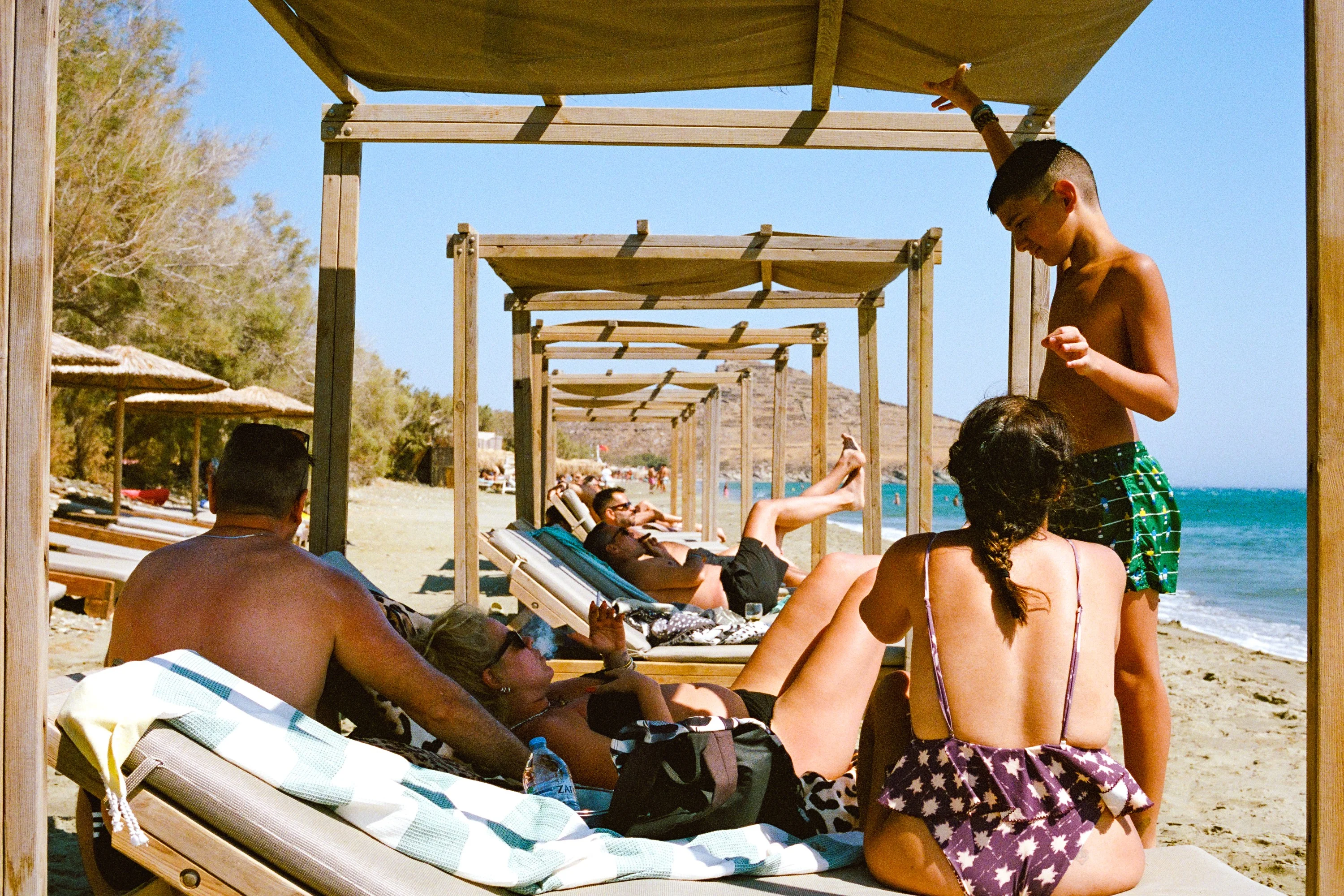 Sunbathers under a row of reed shades, a boy standing over them