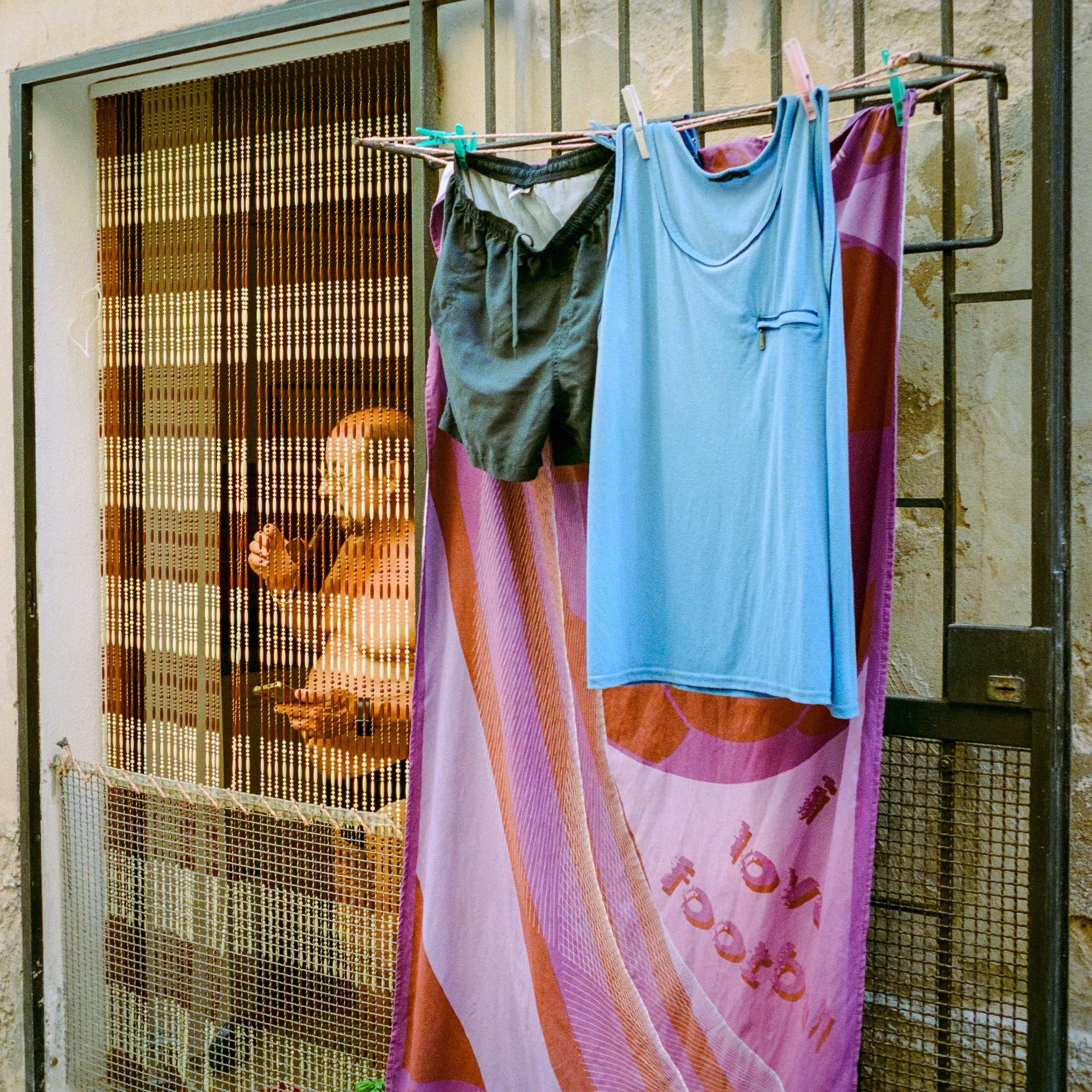 A shirtless man smoking behind a beaded curtain, with shorts, a vest and a towel drying on the window bars