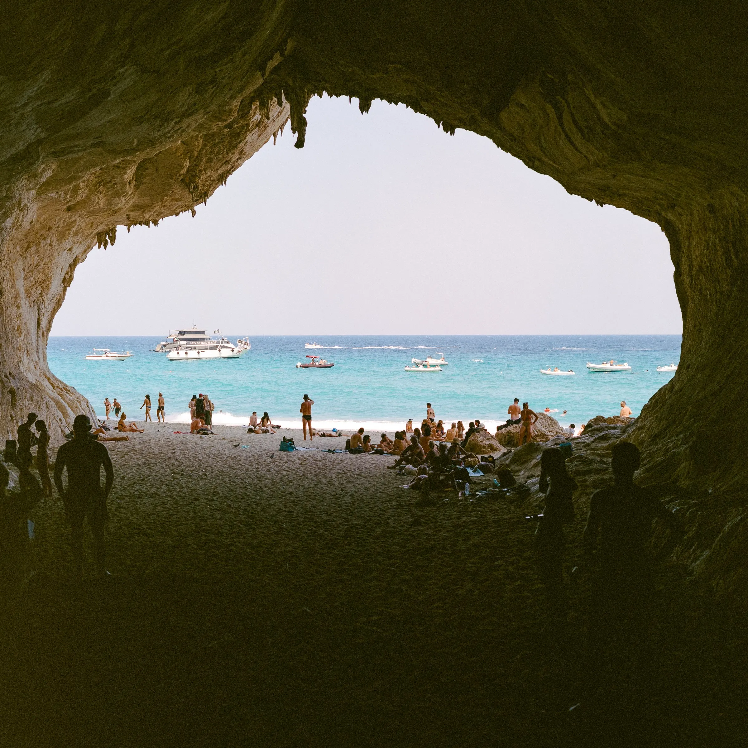 Looking out from a dark sea cave onto a sunlit beach and boats on turquoise water