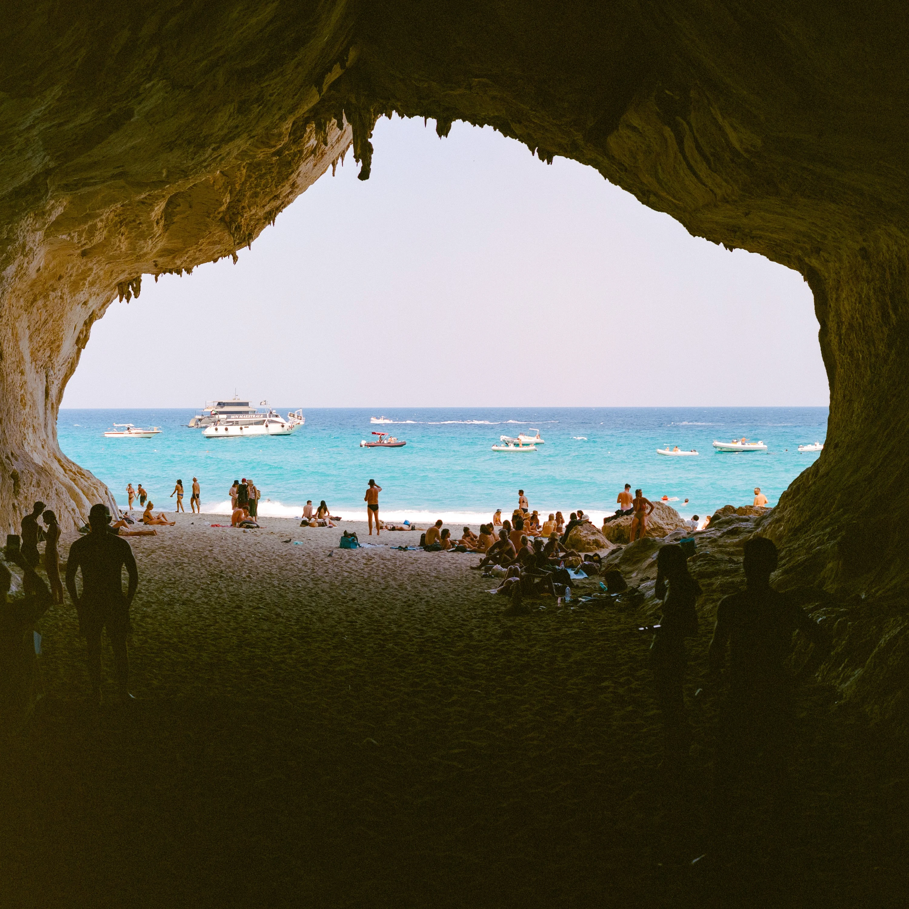Looking out from a dark sea cave onto a sunlit beach and boats on turquoise water