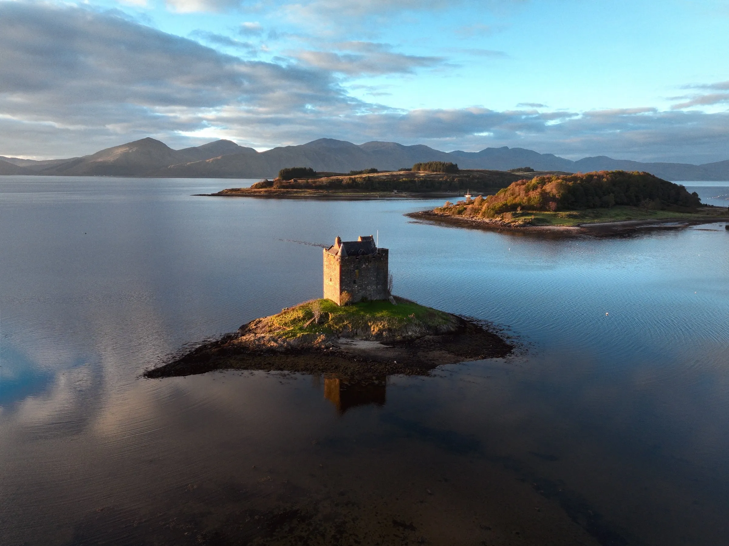 Castle Stalker on its tidal islet in Loch Laich, lit gold at sunrise, with mountains beyond