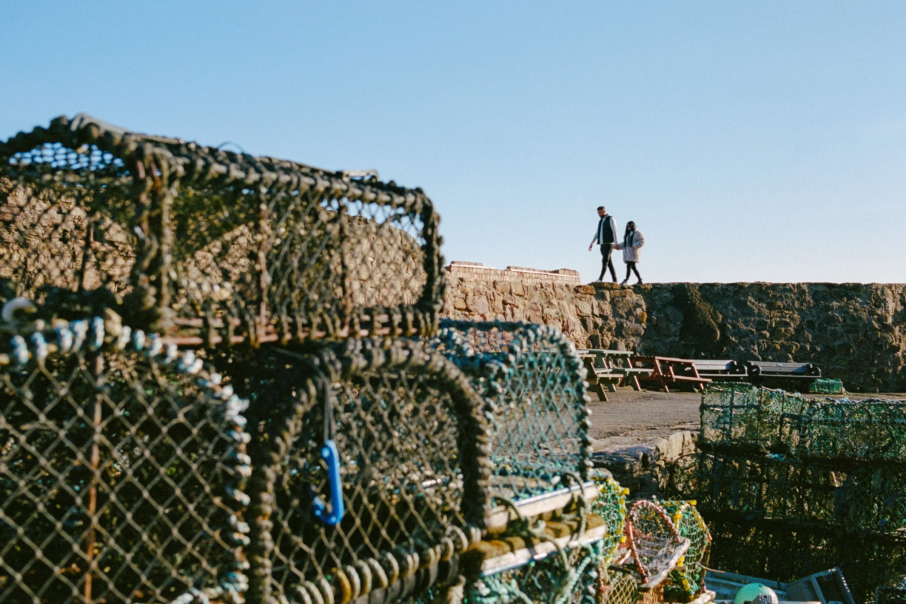 Lobster creels stacked on a harbour wall, two people walking above
