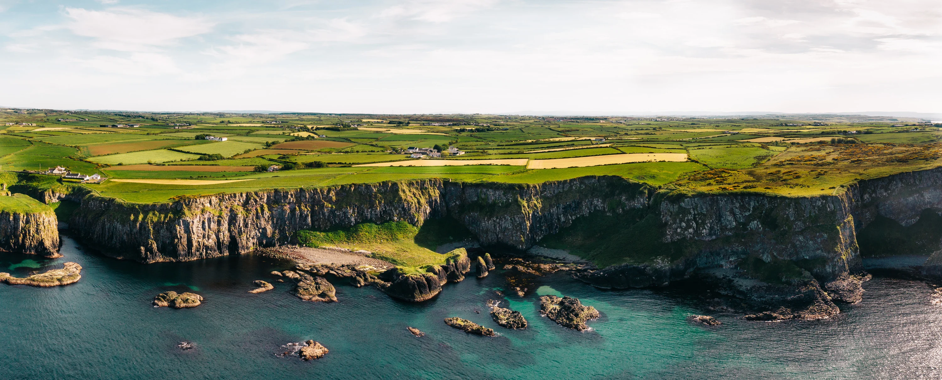 Aerial panorama of sheer basalt cliffs above turquoise water and rocks, a patchwork of green fields stretching inland
