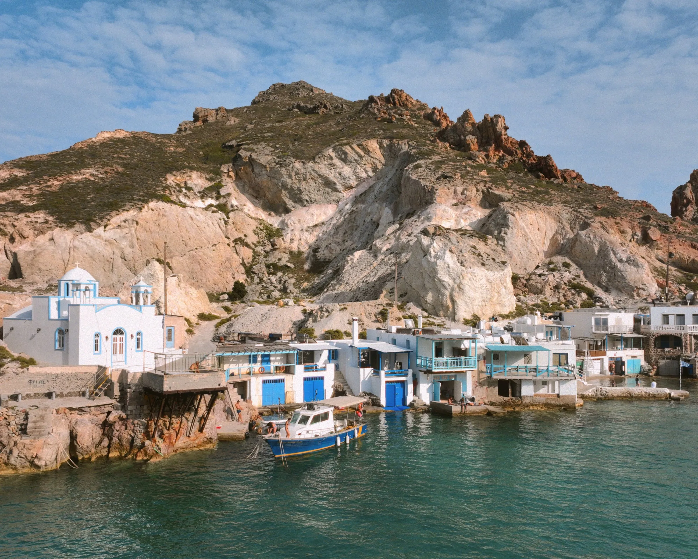 A white chapel and boat-garage houses at the foot of pale cliffs, with a blue fishing boat moored in green water