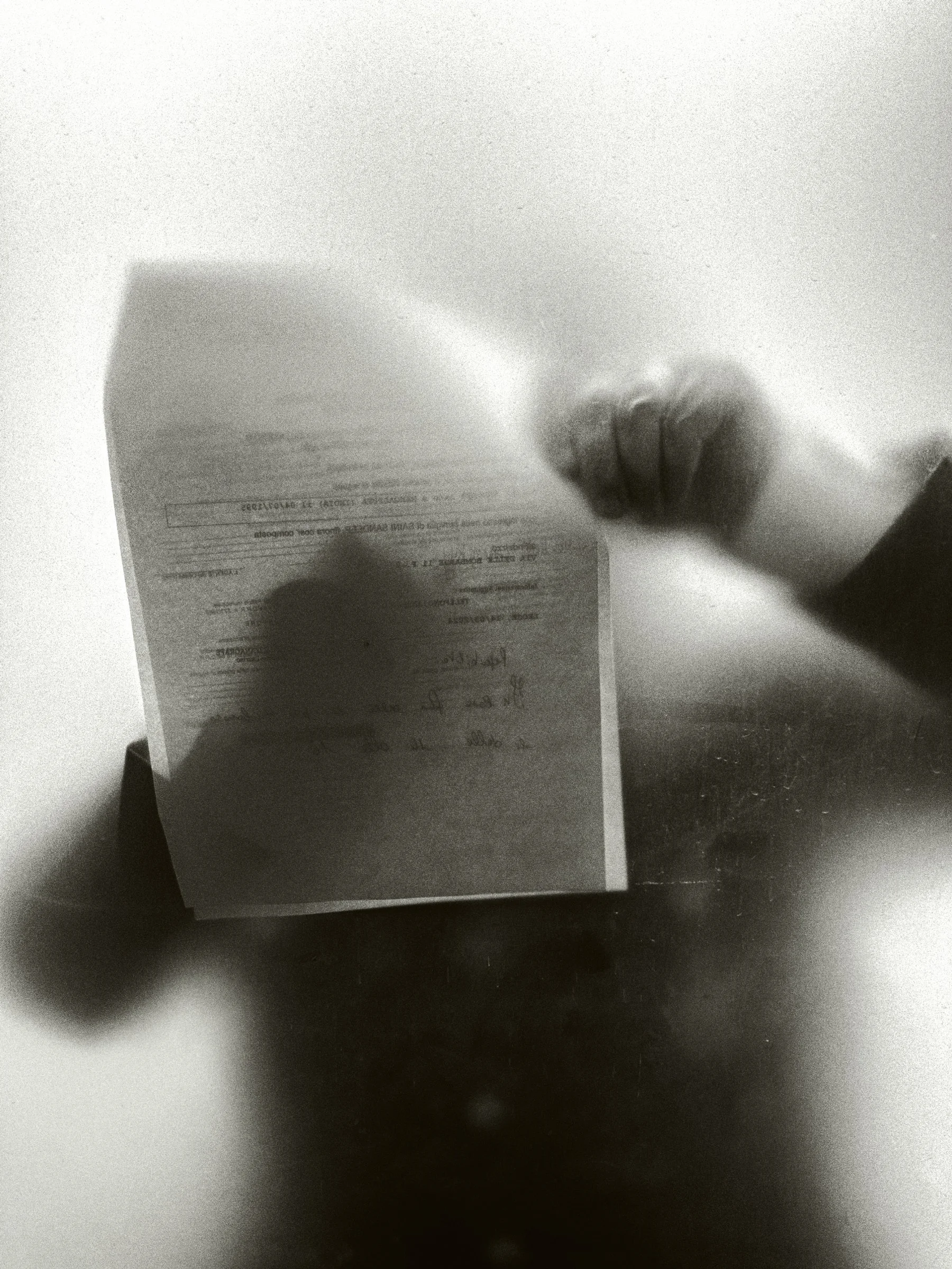 Black and white: a hand presses a sheet of paper against frosted glass, the figure behind it a soft shadow