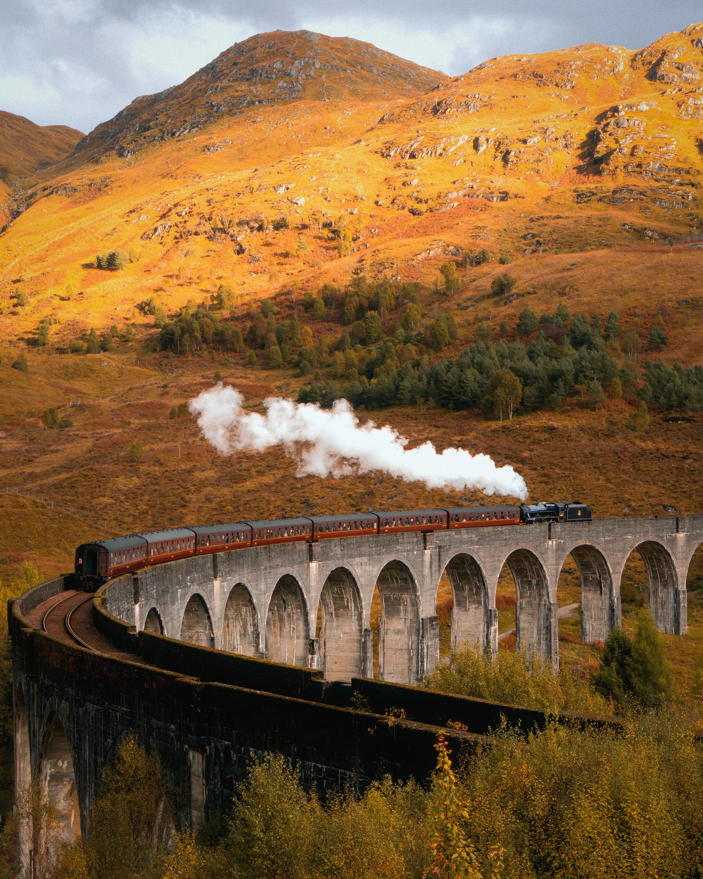 A steam train crossing the curved Glenfinnan viaduct below golden hills