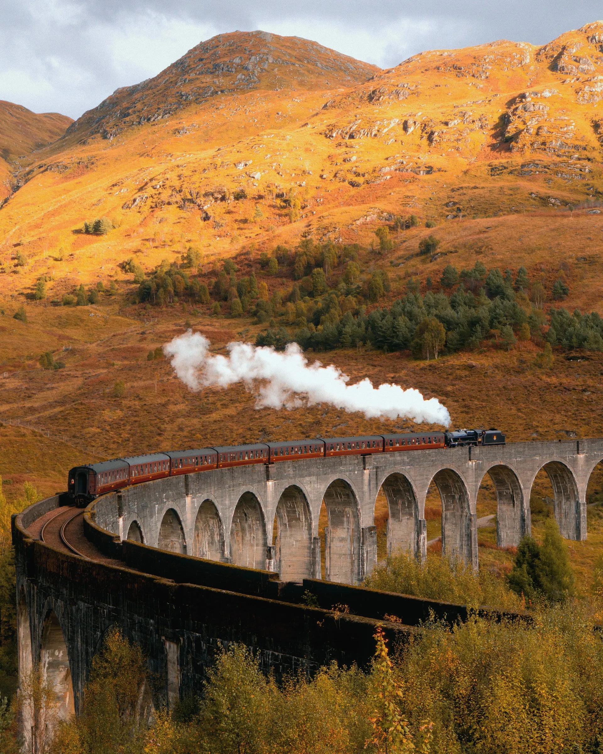 A steam train crossing the curved Glenfinnan viaduct below golden hills