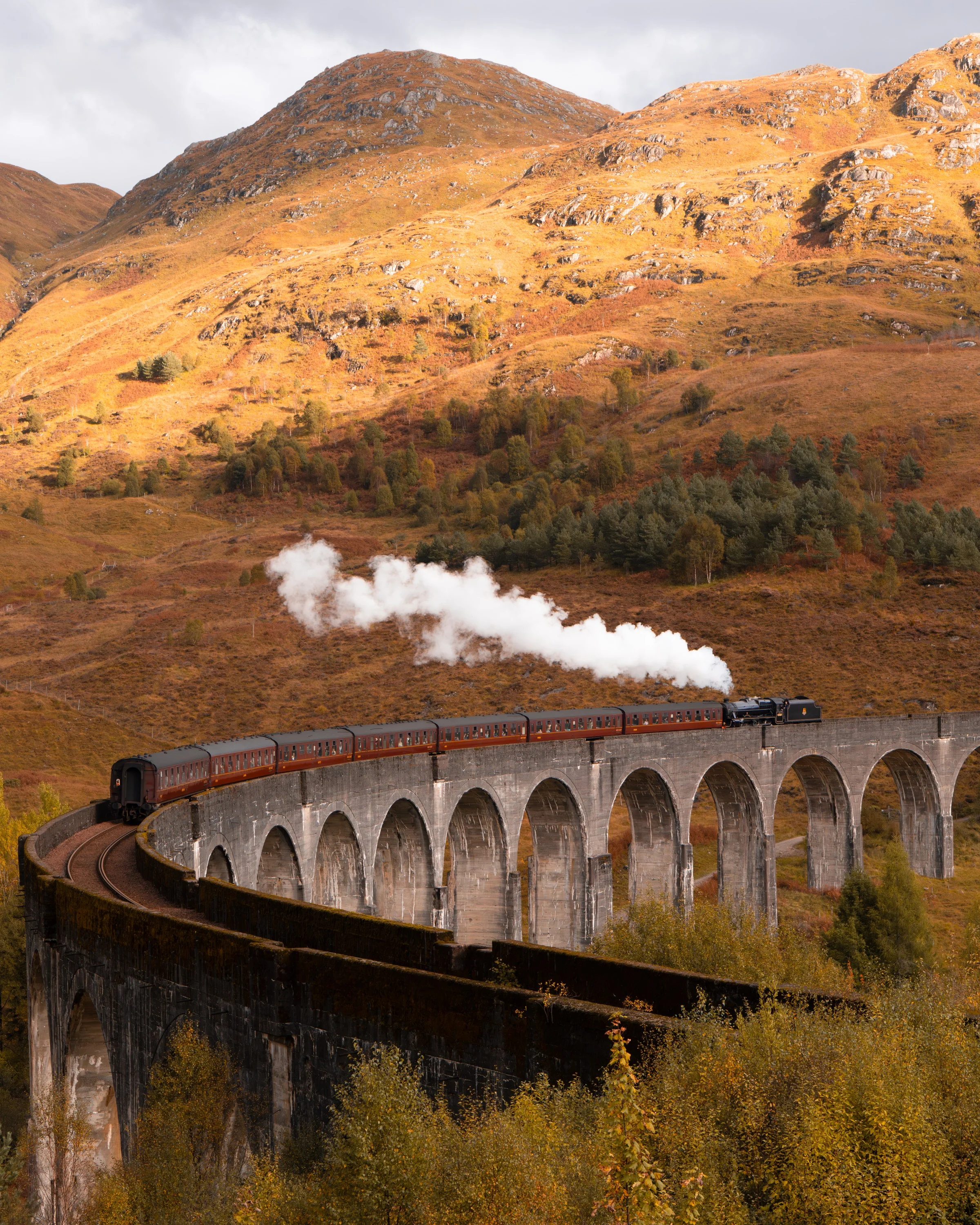 A steam train crossing the curved Glenfinnan viaduct below golden hills