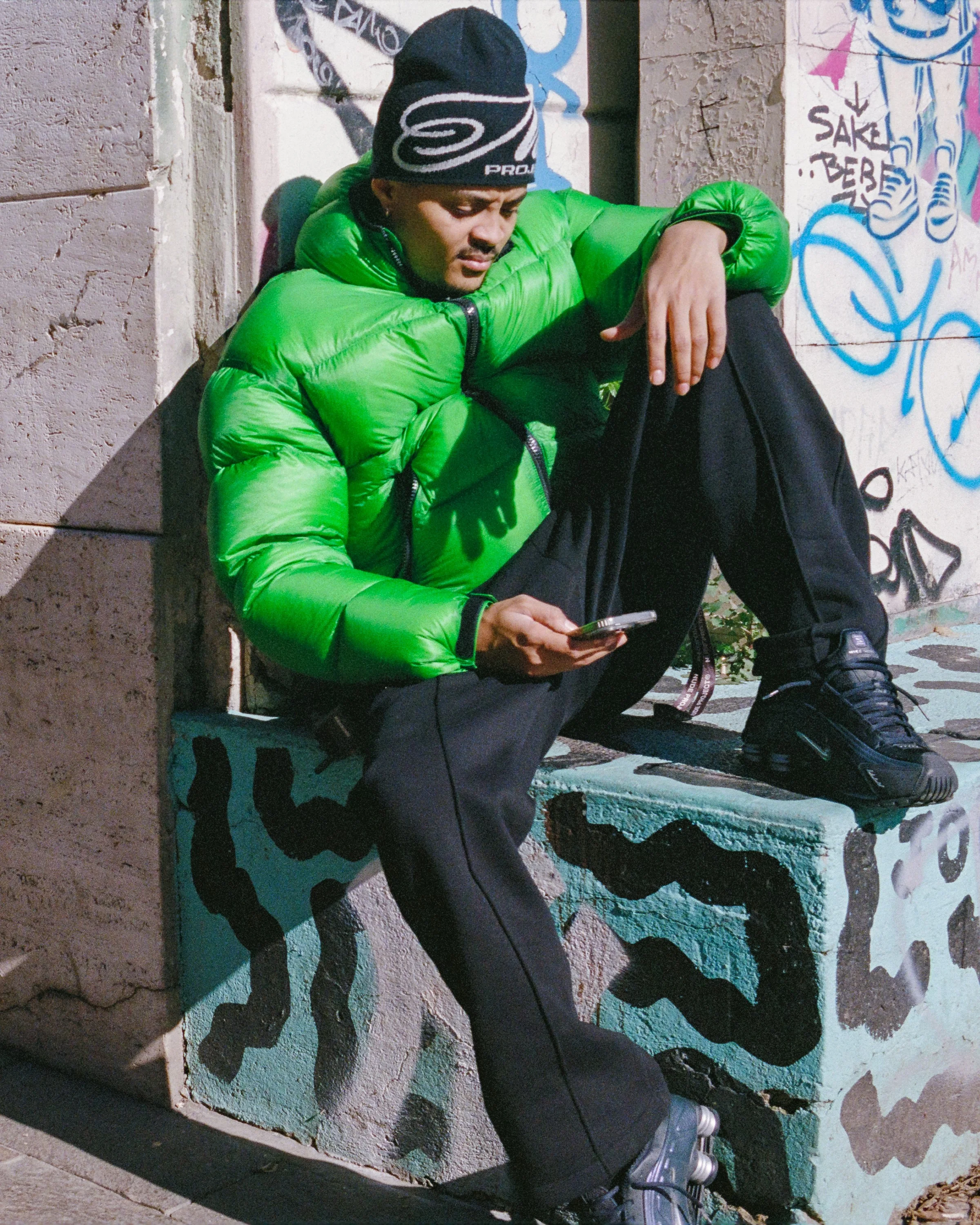 A man in a bright green puffer jacket sitting by a graffitied wall