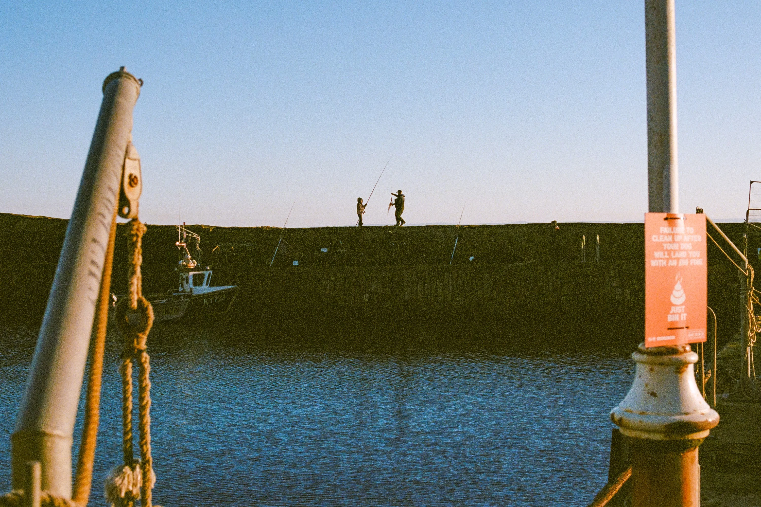 A harbour wall at dusk, two figures fishing at the end