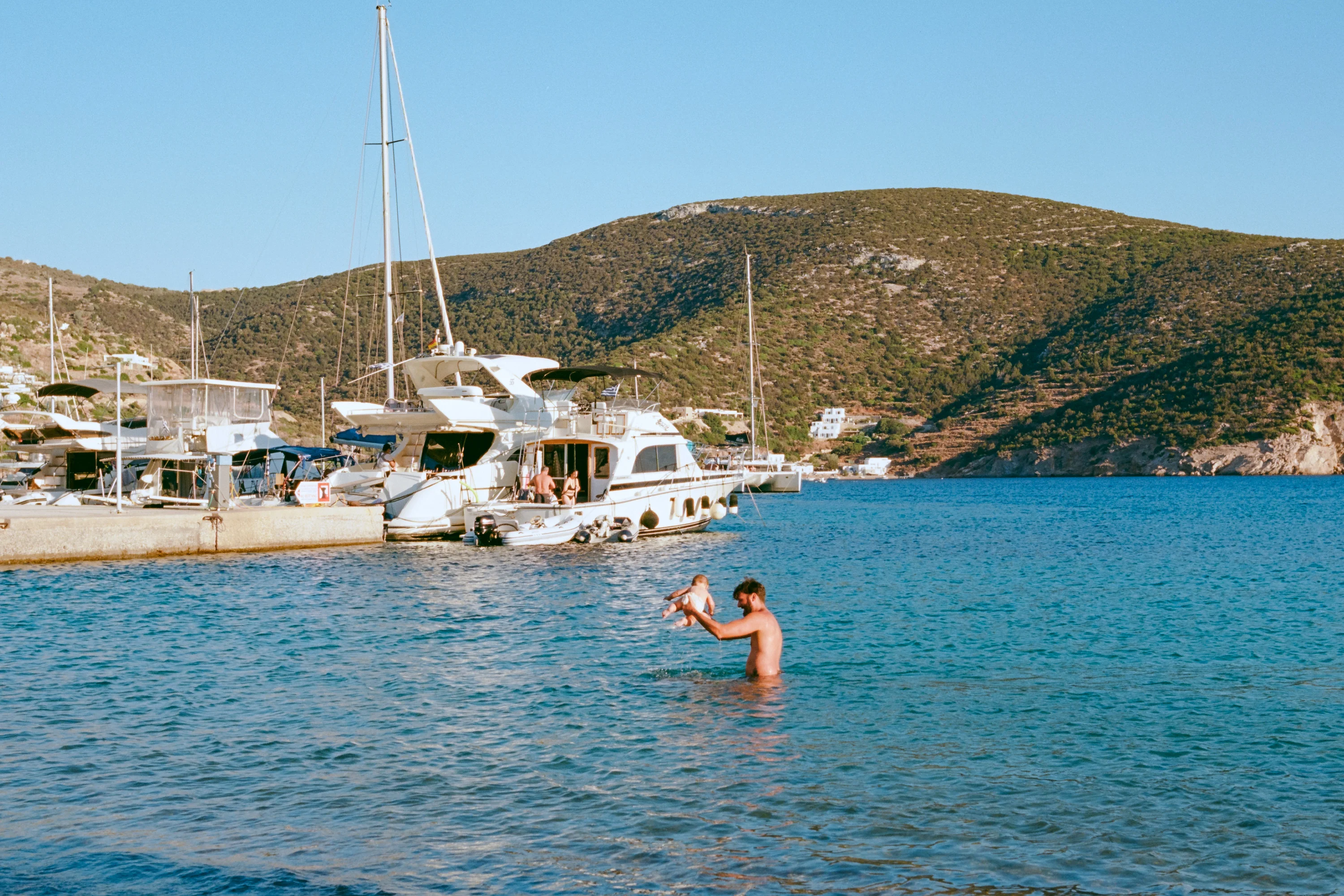 A man wading in a calm bay in front of moored yachts and a dry green hillside