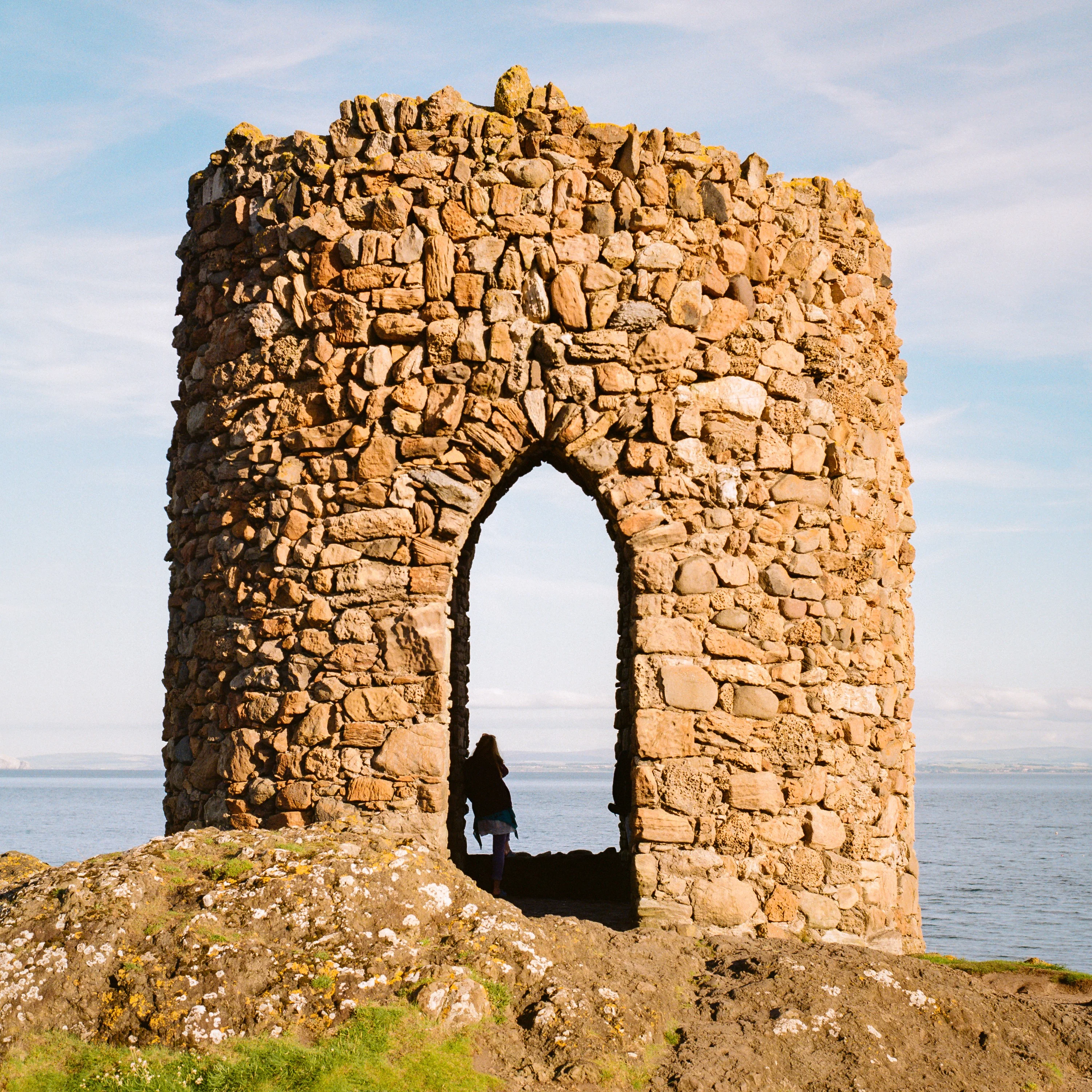 A figure stands in the arched doorway of a ruined stone tower by the sea