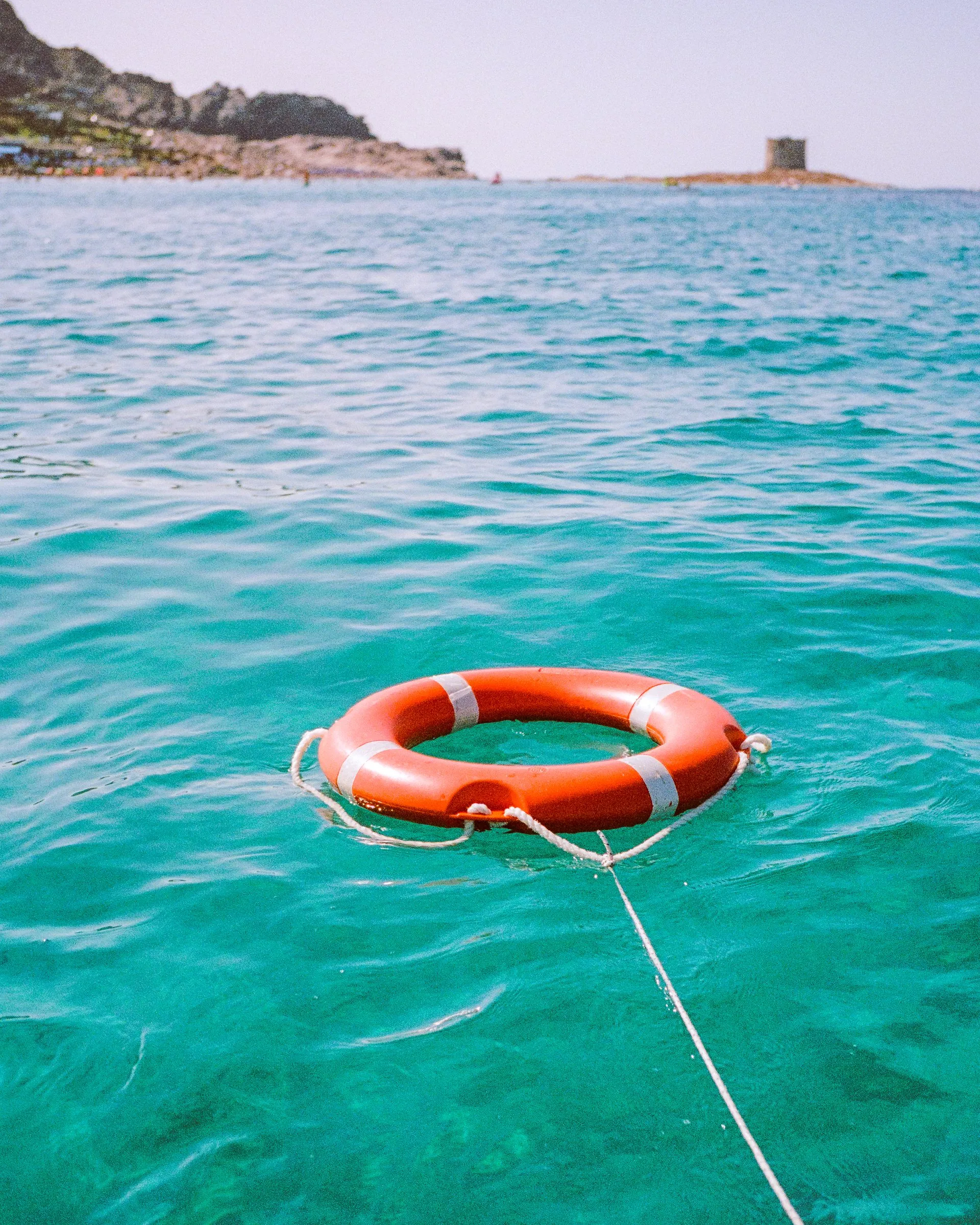 An orange lifebuoy floating on clear turquoise water, a watchtower on the horizon