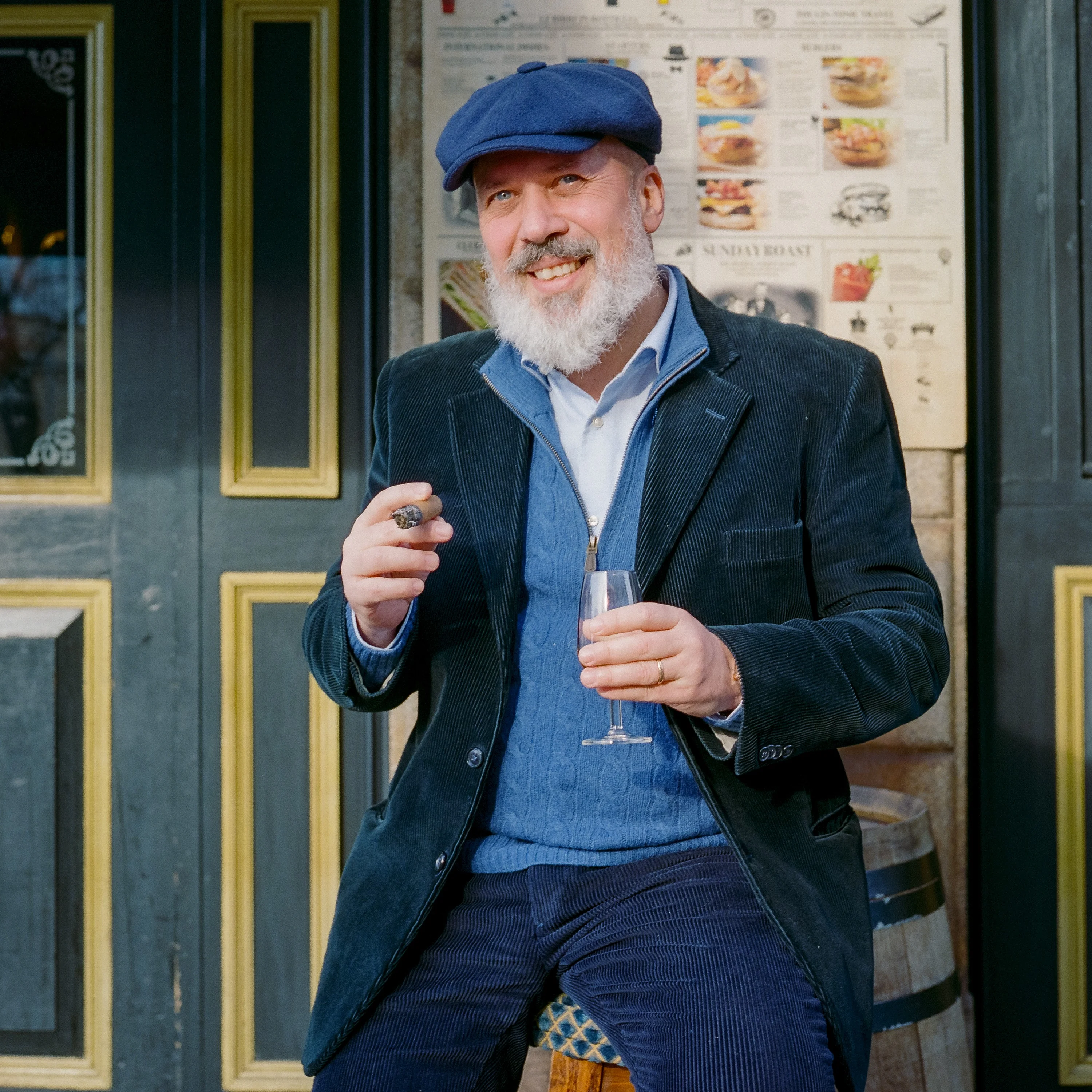 A bearded man in a flat cap with a cigar and a glass