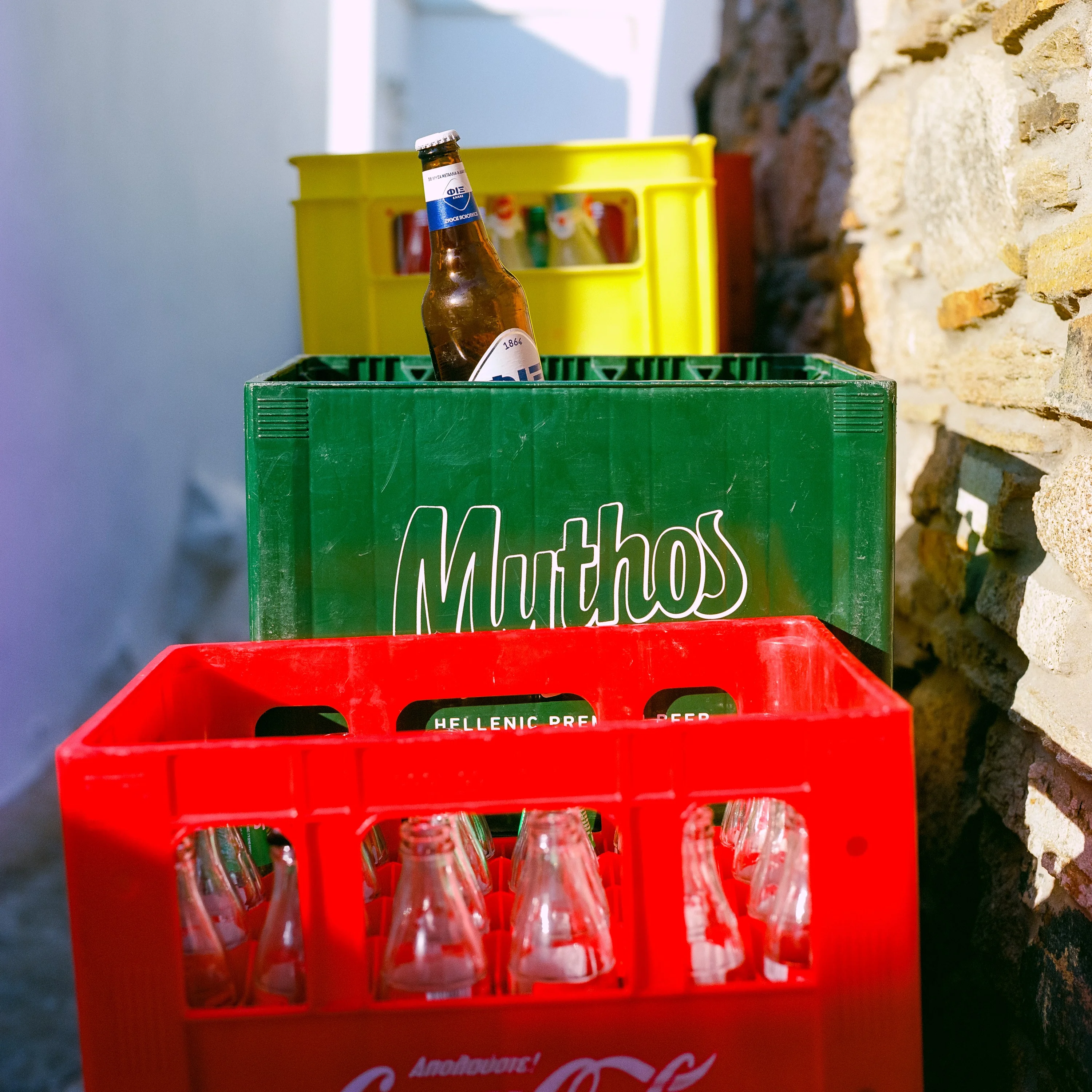 Red, green and yellow bottle crates stacked in a whitewashed alley, a beer bottle on top