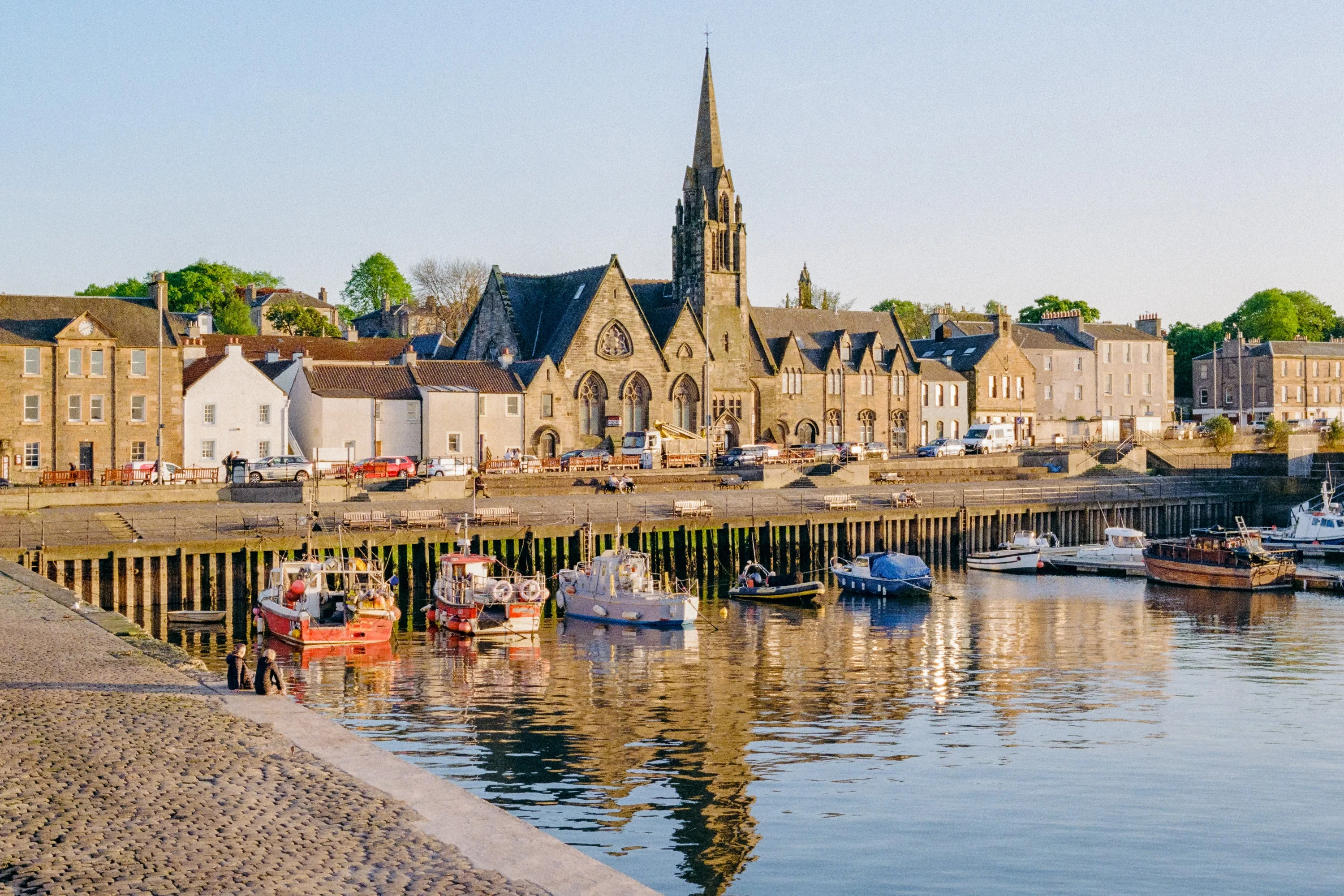 Fishing boats in Newhaven harbour below a church spire and stone houses in evening light