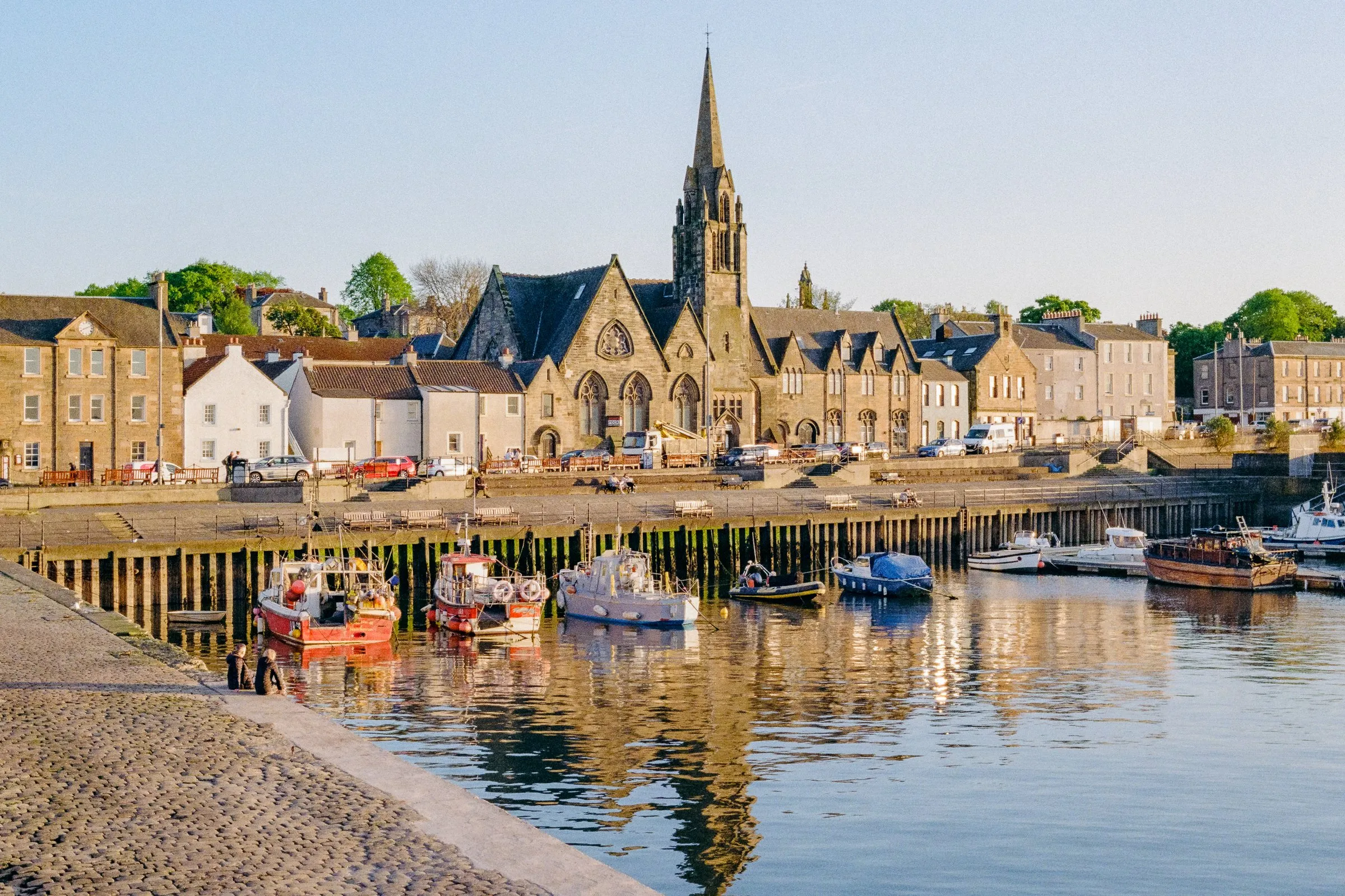Fishing boats in Newhaven harbour below a church spire and stone houses in evening light