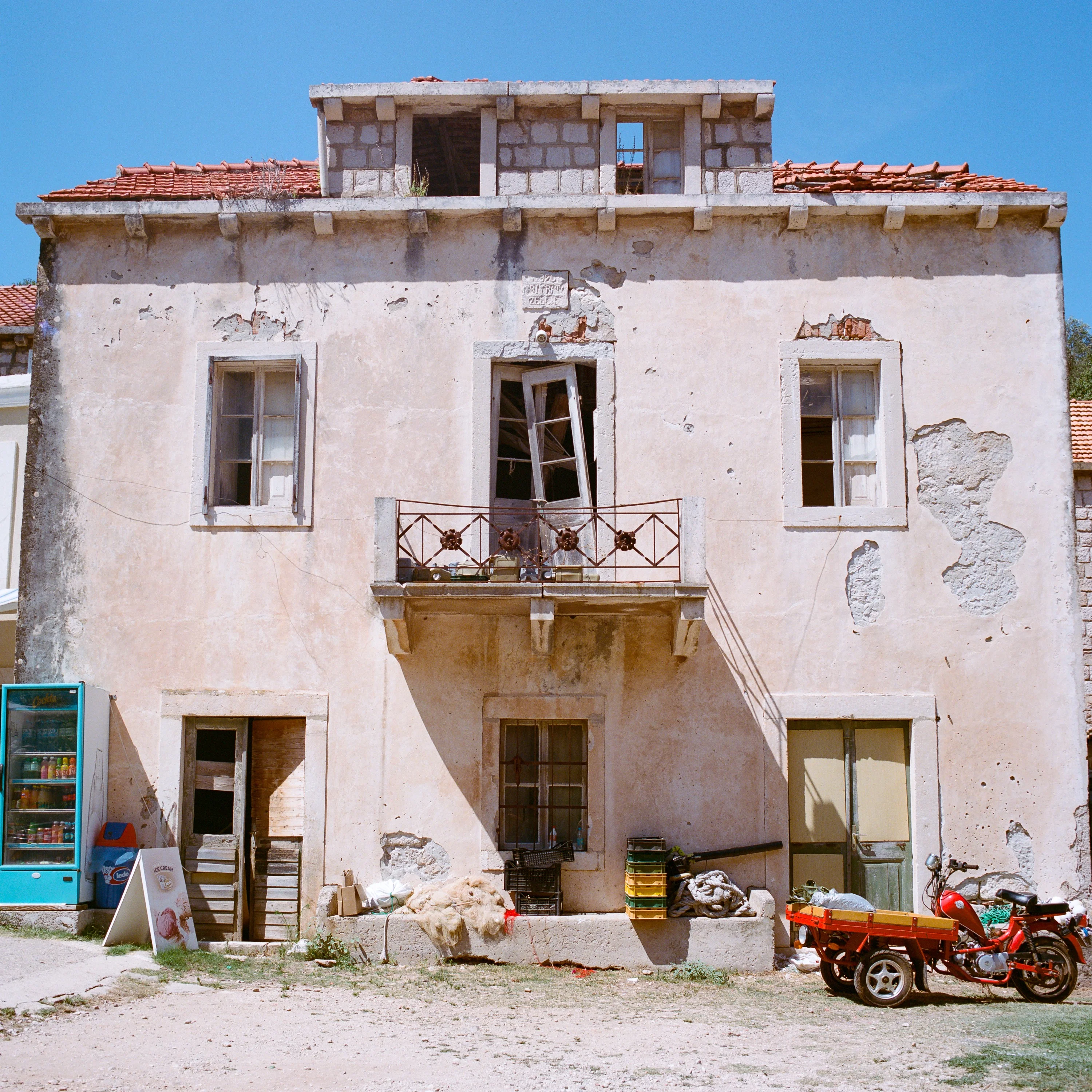 A crumbling stone house with a balcony, a red three-wheeler parked in front