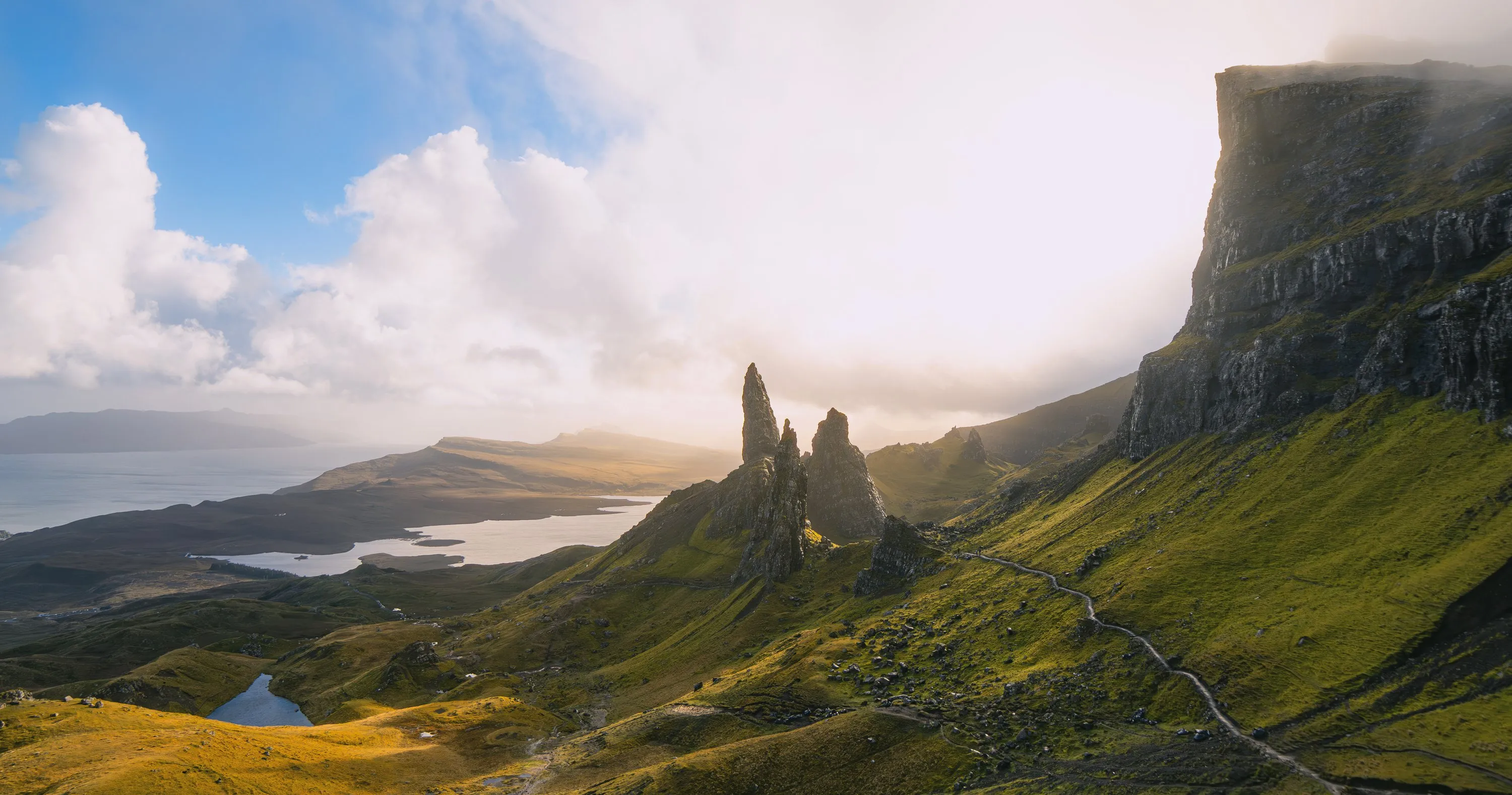 The Old Man of Storr's rock pinnacles in low sun, with green slopes, a winding path and lochs below a bright, misty sky