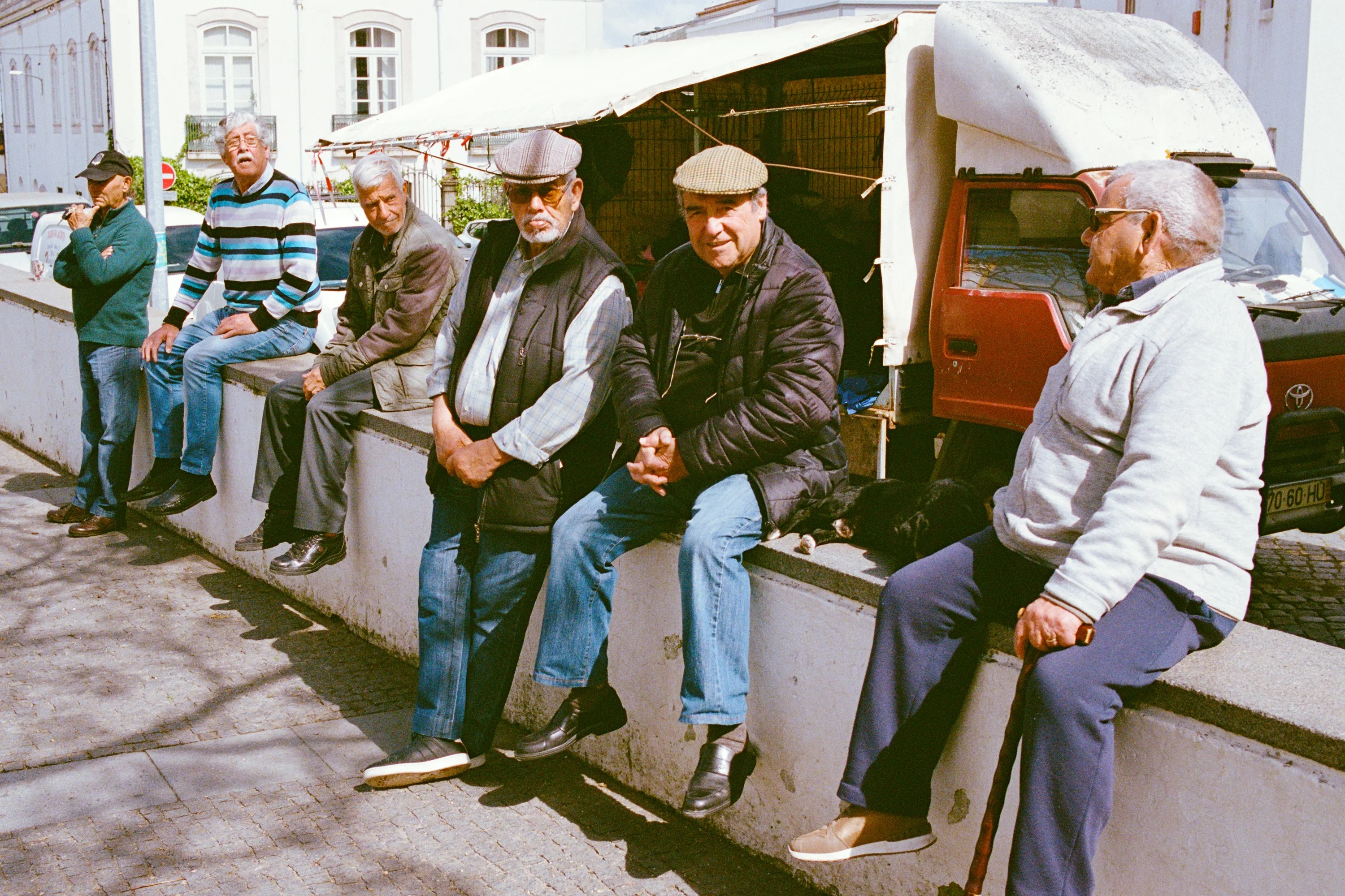 A row of old men sitting on a wall in the sun