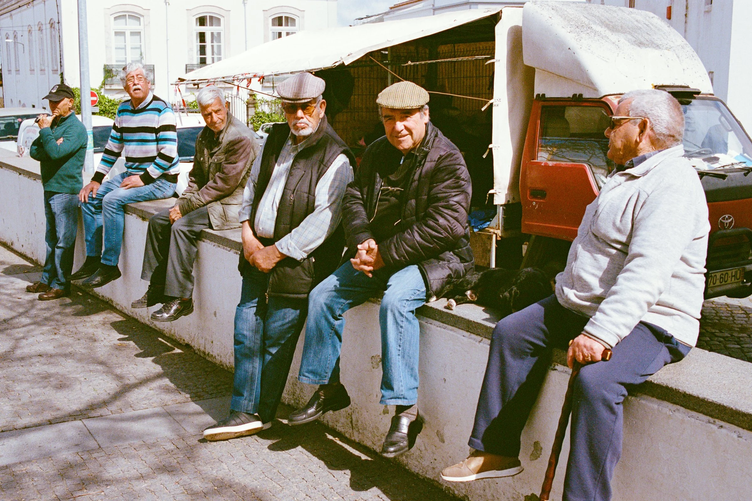 A row of old men sitting on a wall in the sun