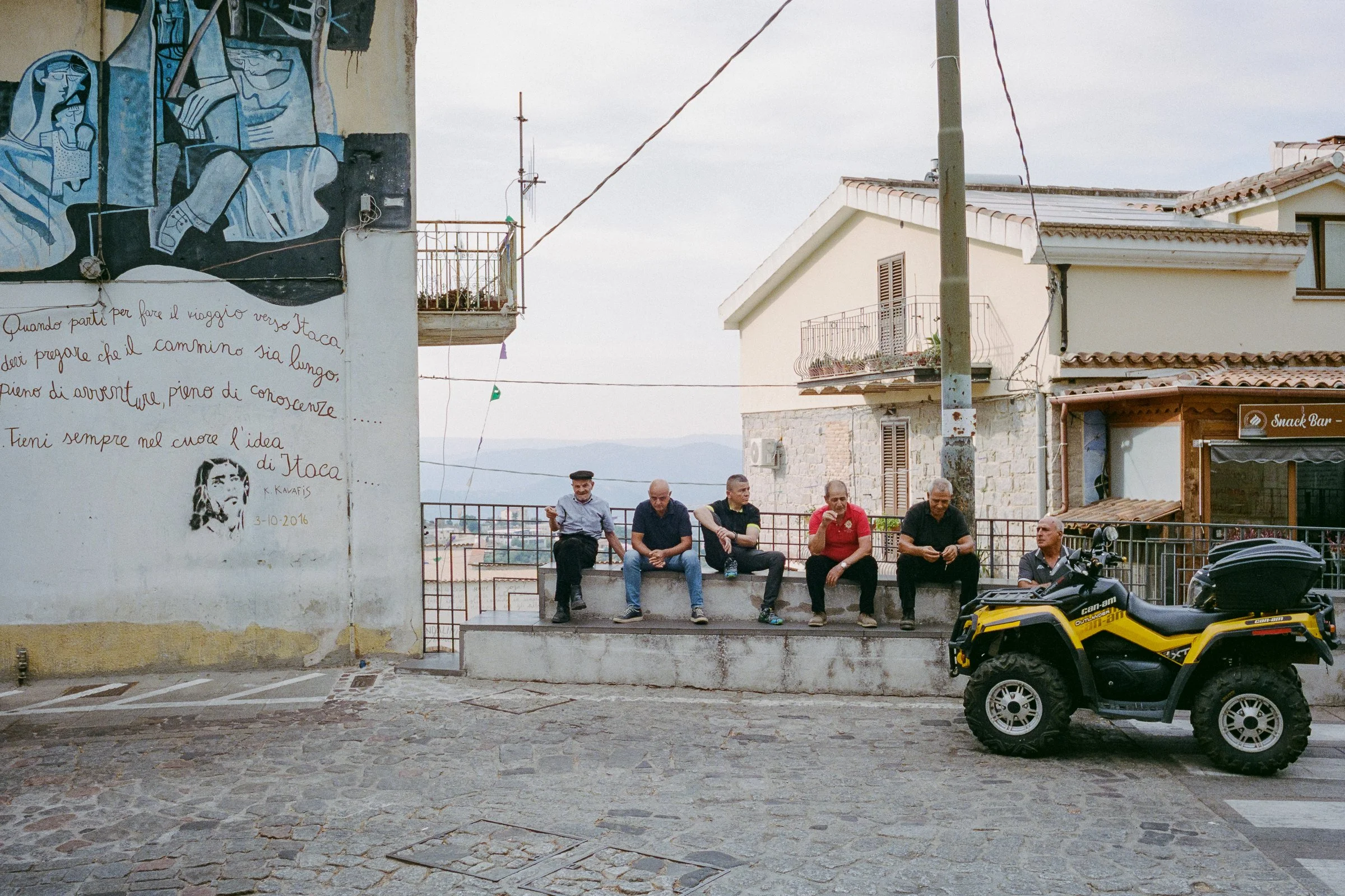 Men sit along a wall beneath a painted mural, a yellow quad bike parked beside them
