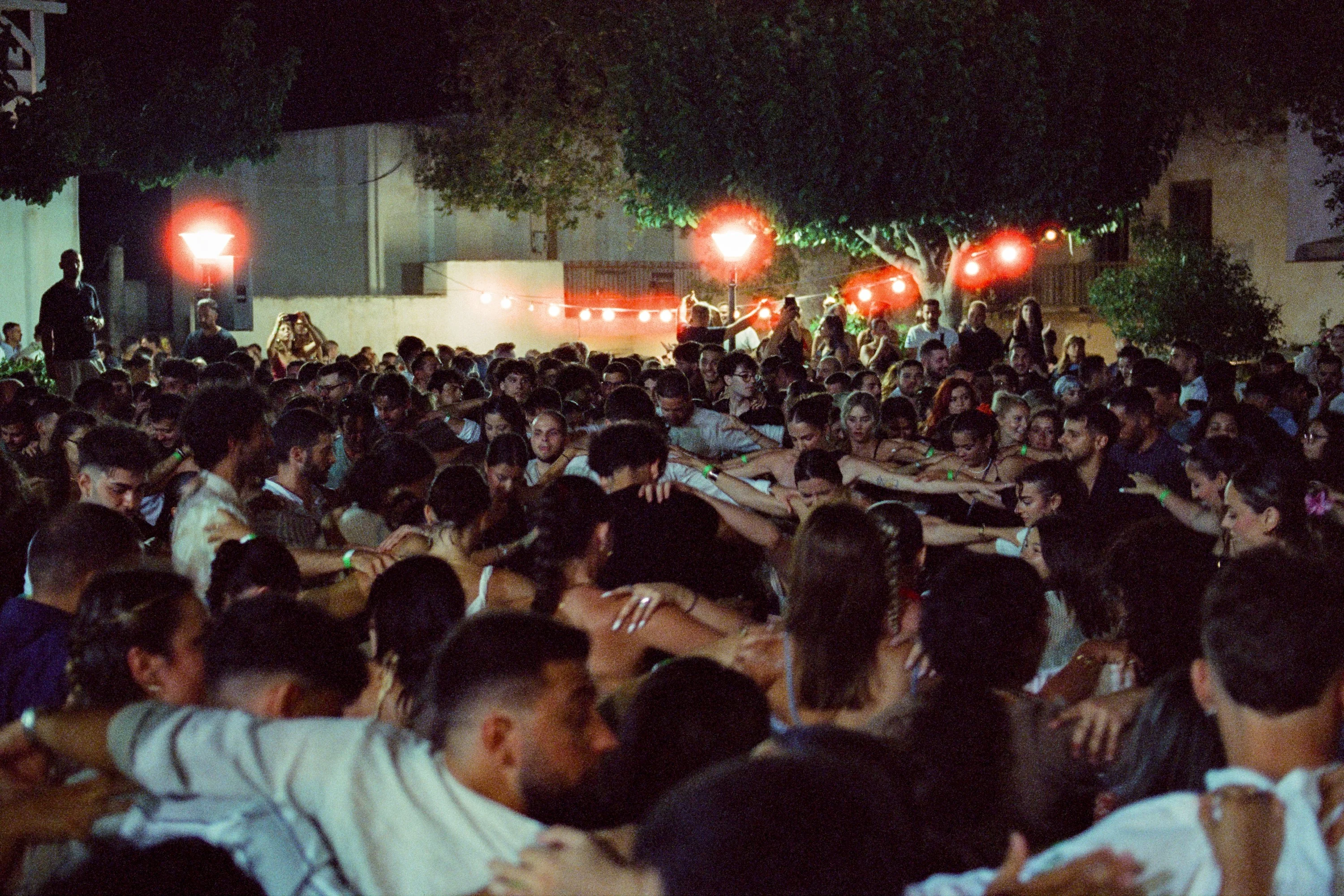 A packed square of dancers under red lights at a village festival