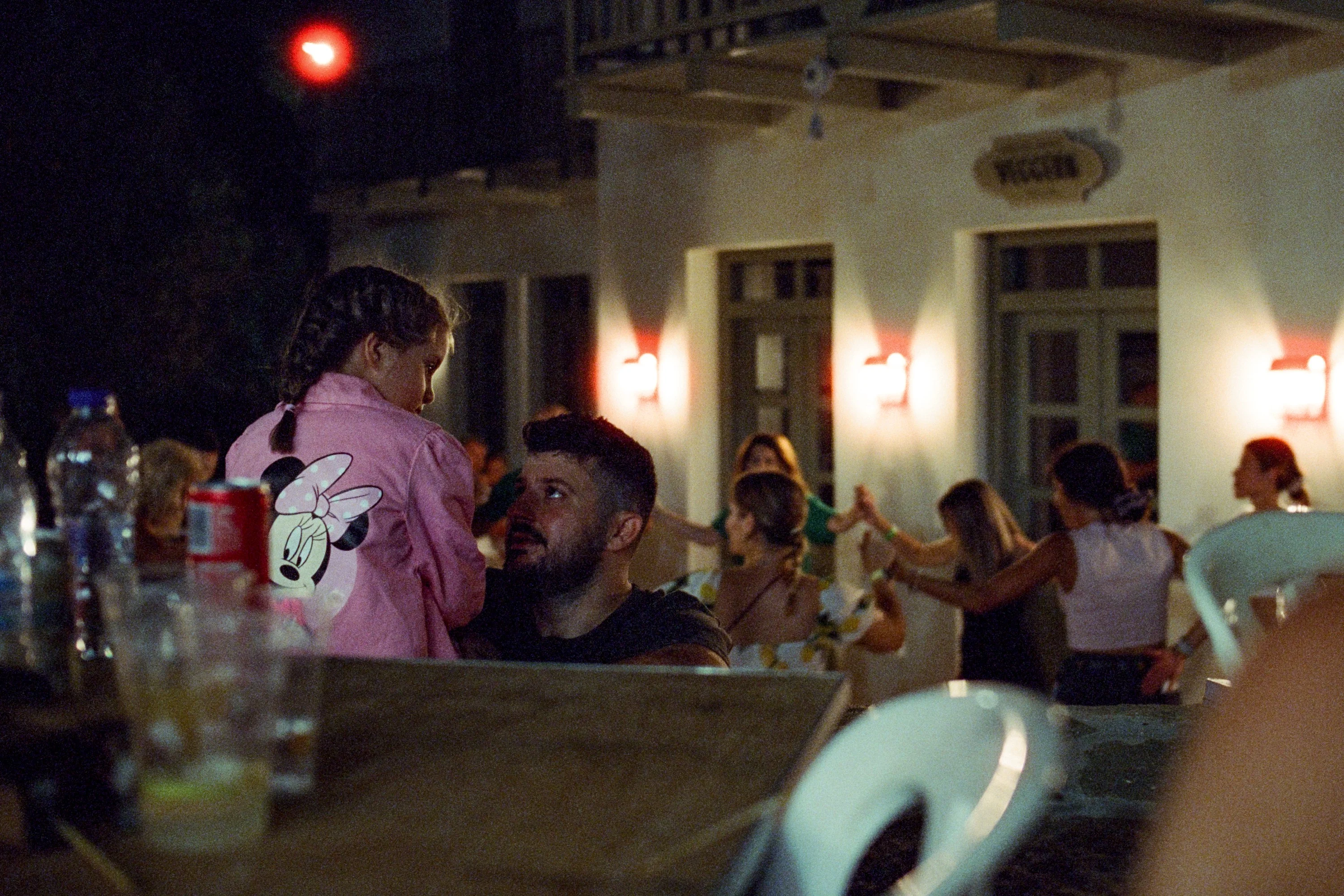 People talking at a table at a night festival, a woman in pink dancing behind
