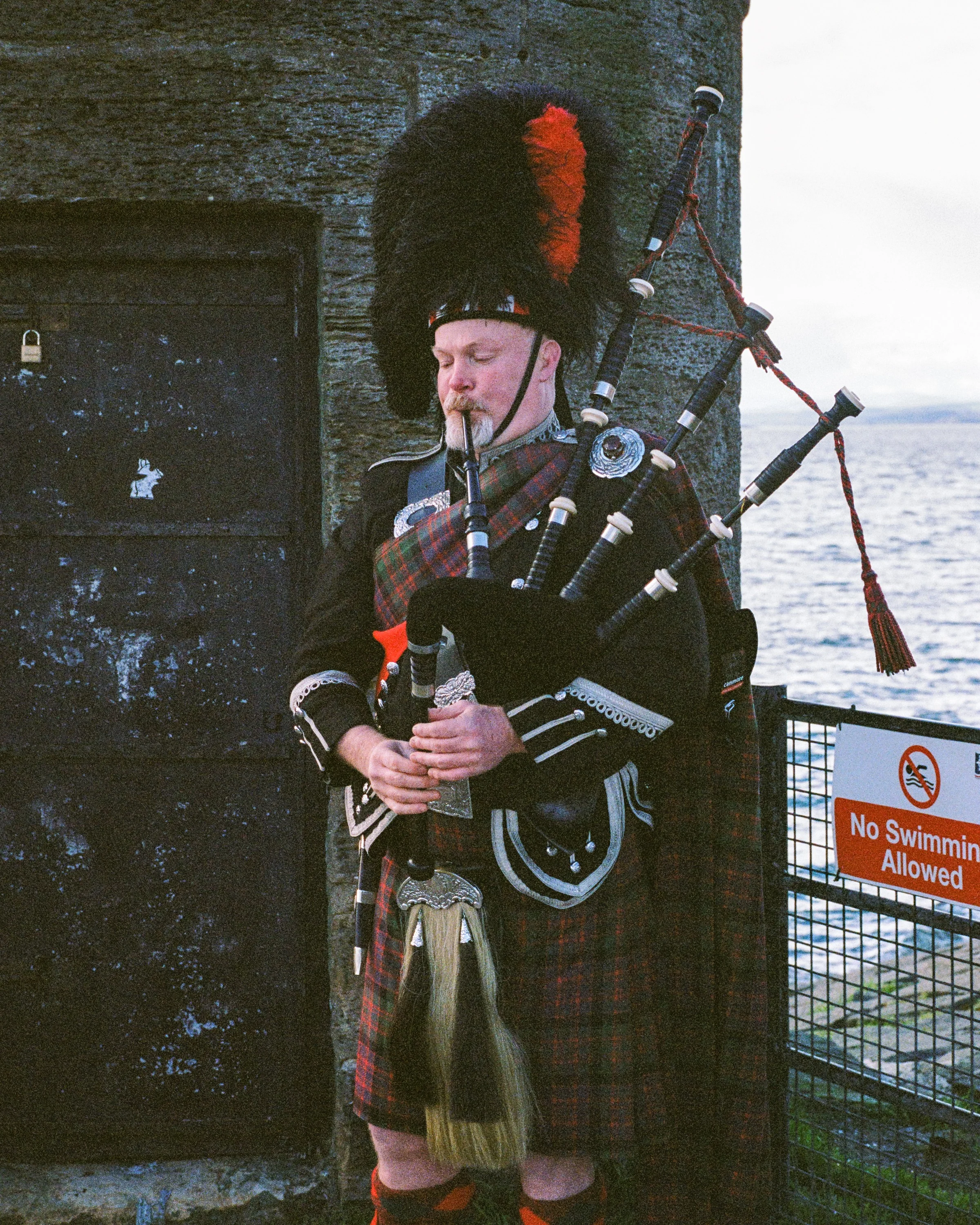 A piper in full dress playing beside a harbour wall