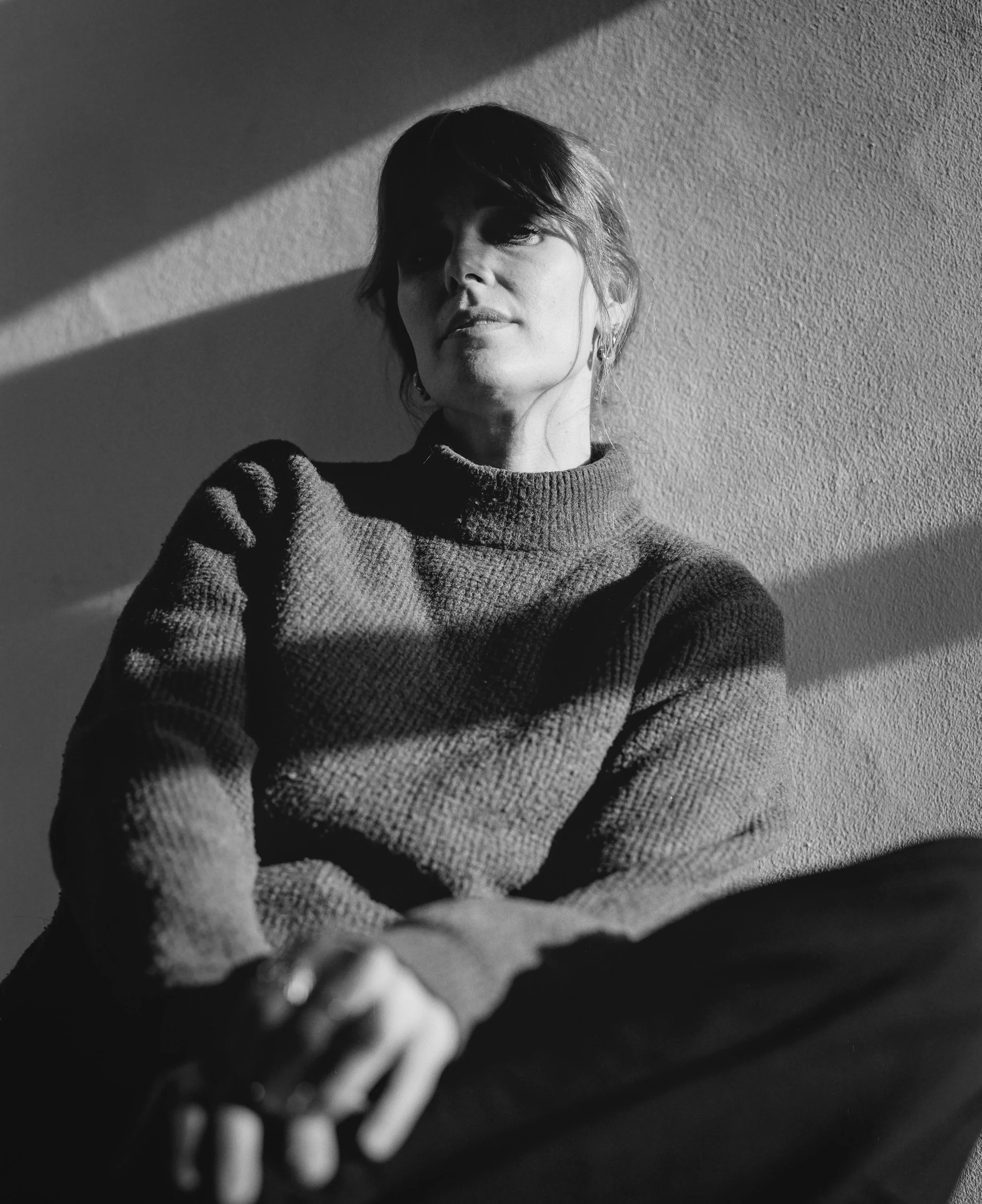 Black and white portrait of a woman in a dark jumper sitting against a sunlit wall
