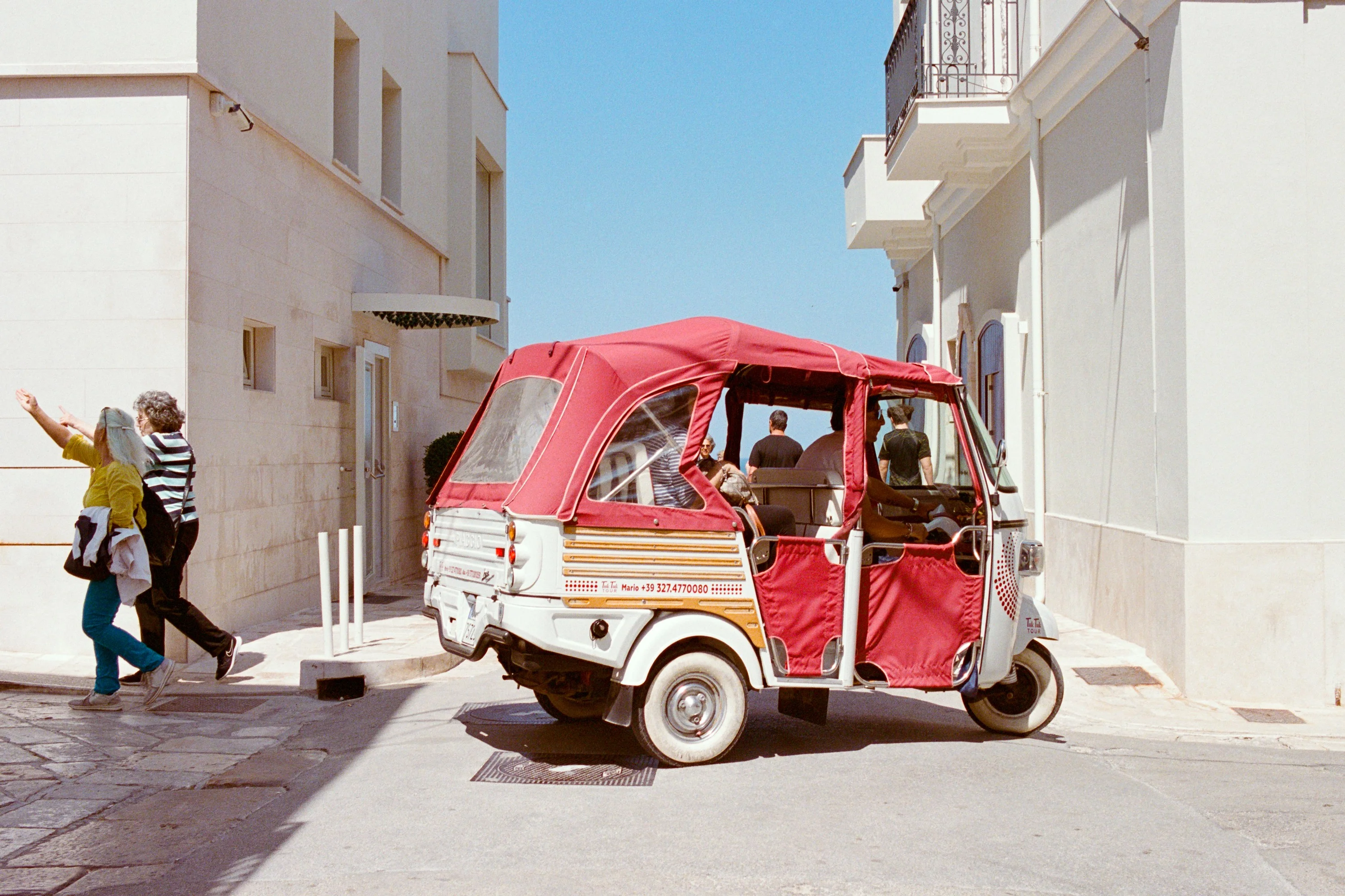 A red and white Ape tuk-tuk turns through a white street as two women walk past