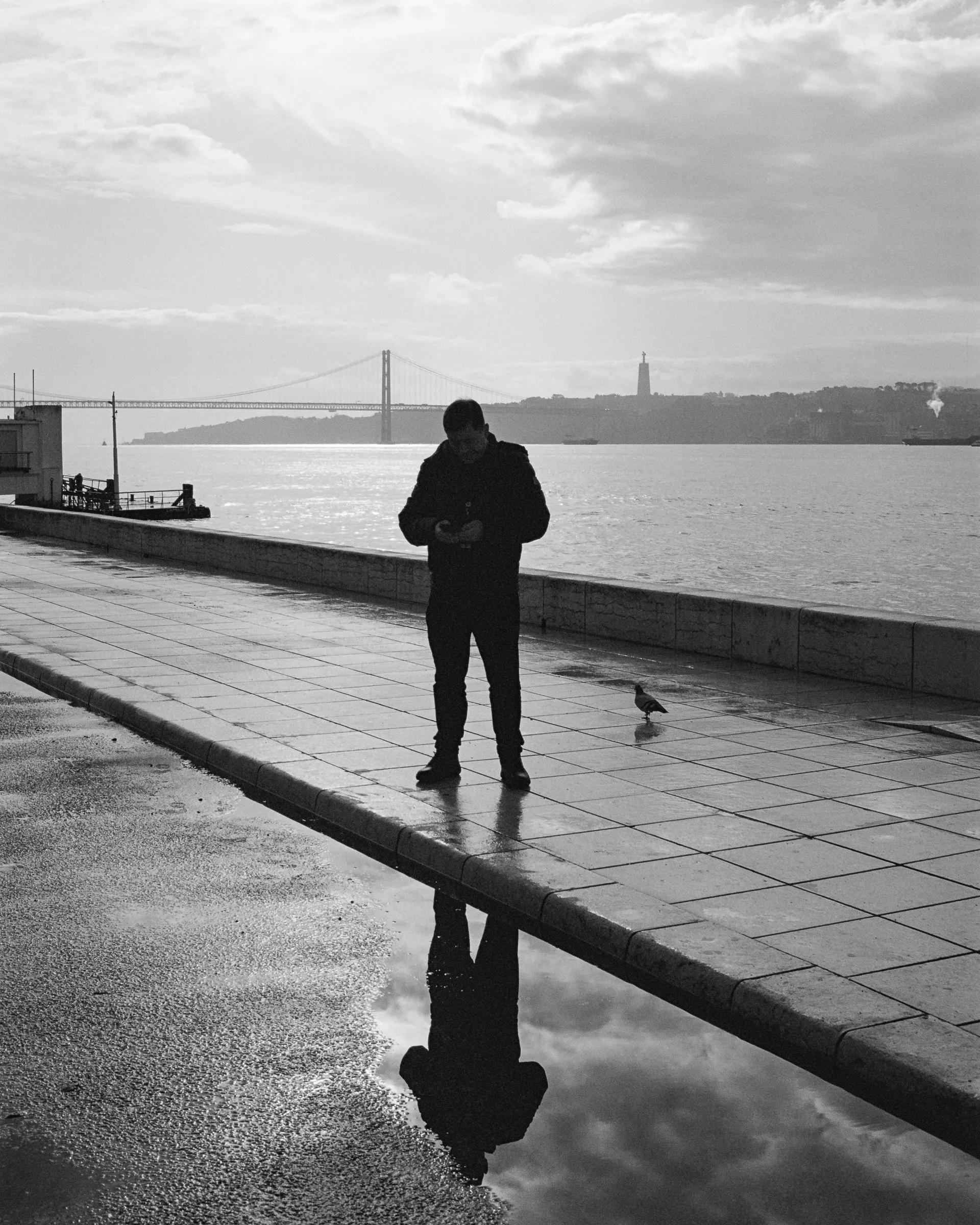 Black and white photo of a man and his reflection on a wet riverside walkway