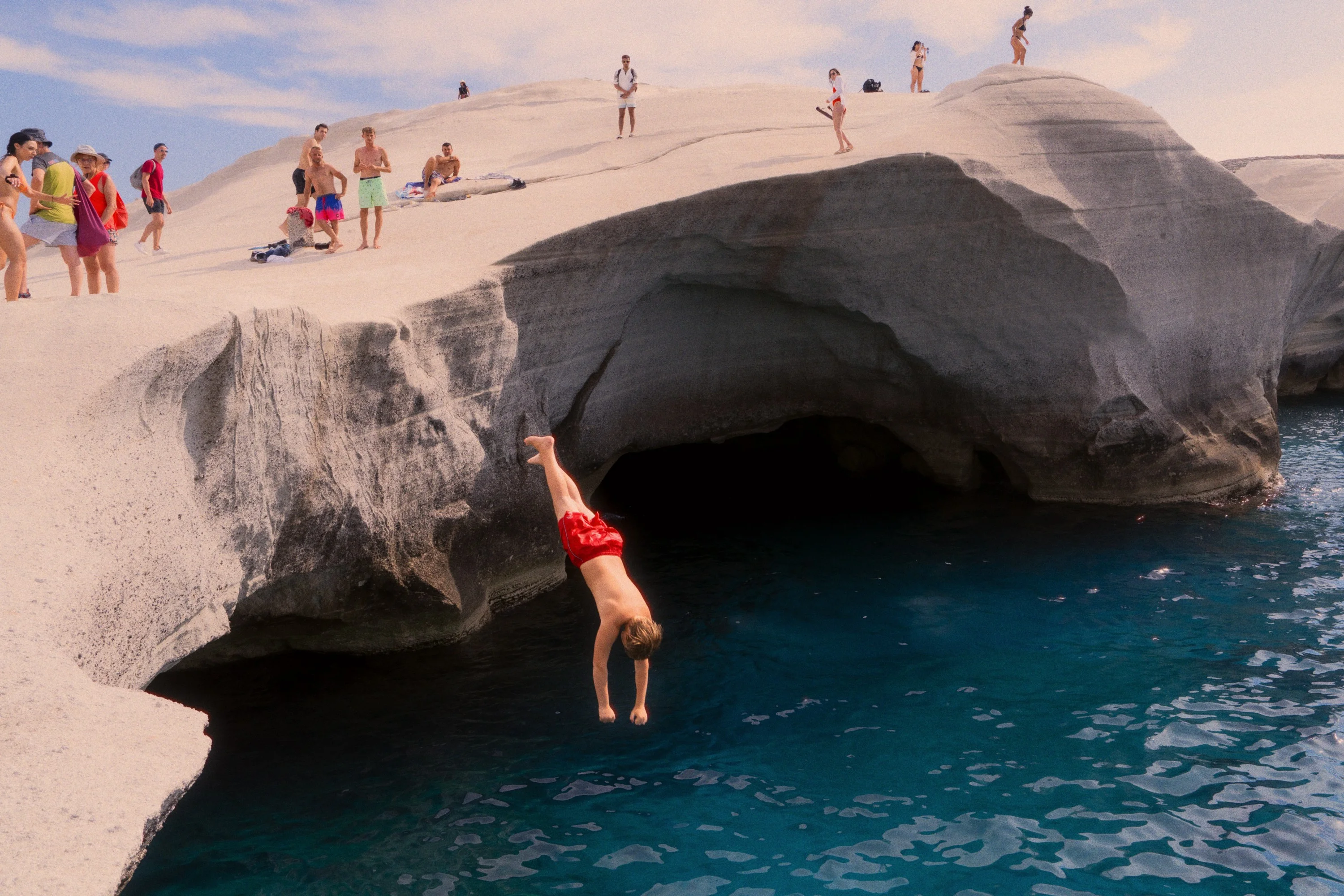 A swimmer in red shorts drops from a white rock arch into dark blue water while people watch from the top