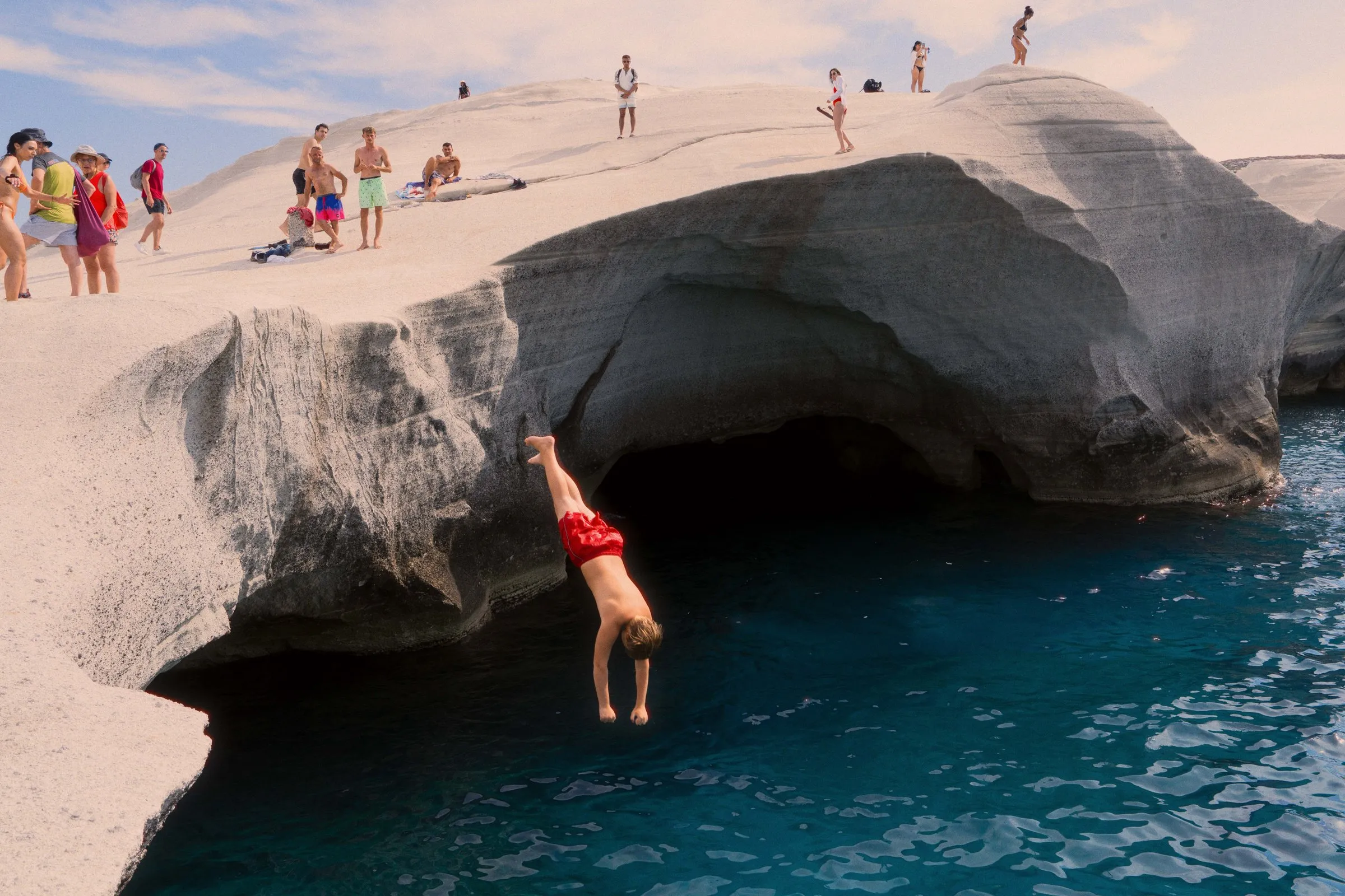A swimmer in red shorts drops from a white rock arch into dark blue water while people watch from the top