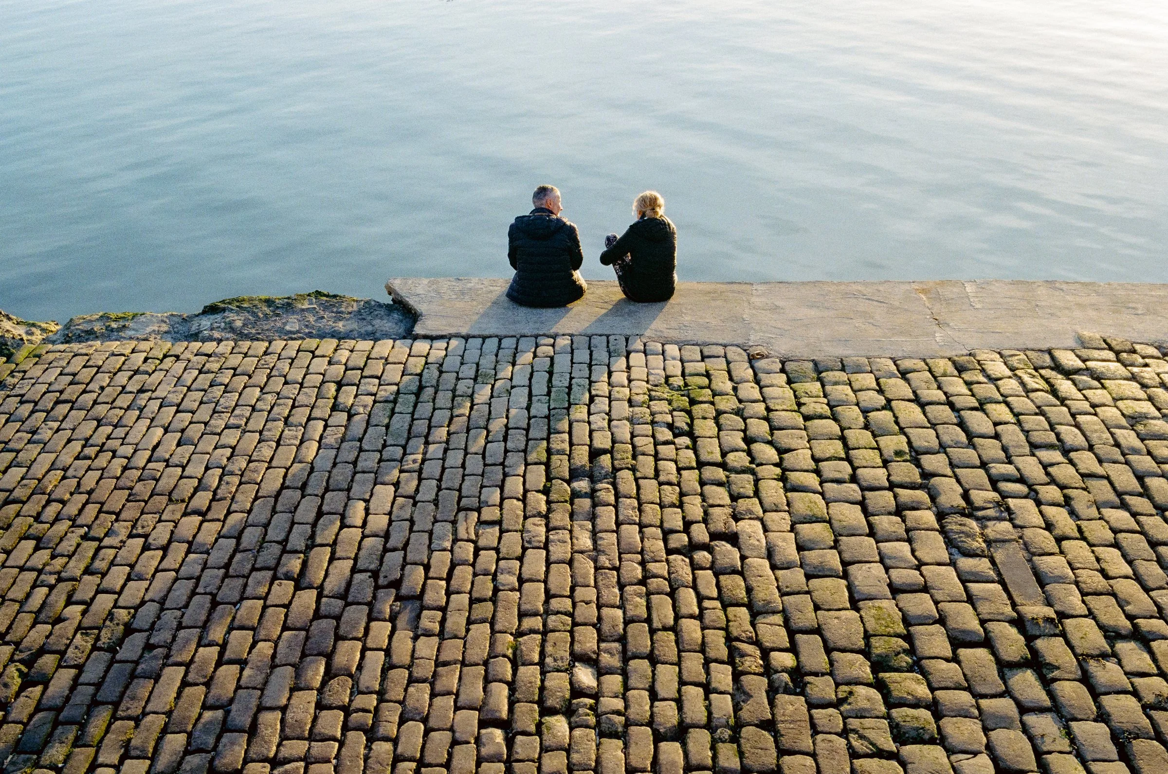 Two people sit at the edge of a cobbled slipway looking out over calm water
