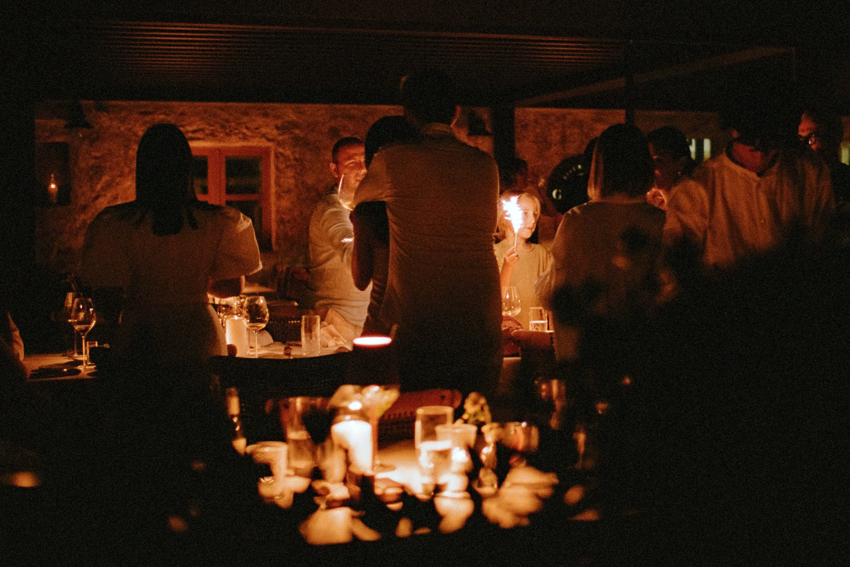 A candlelit dinner on a stone terrace at night, a child holding a sparkler among the guests