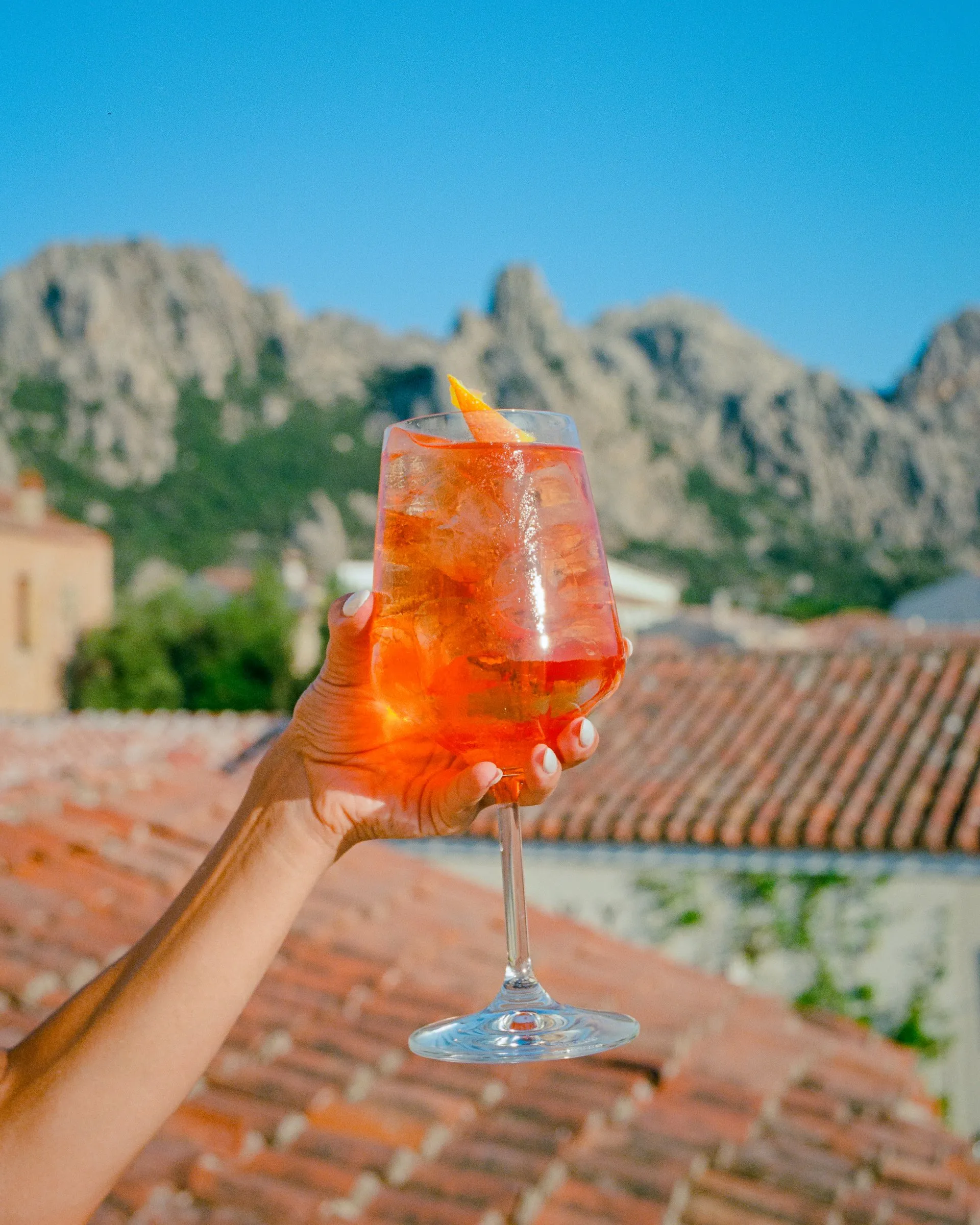 A hand holds up a spritz above terracotta roofs, granite peaks behind