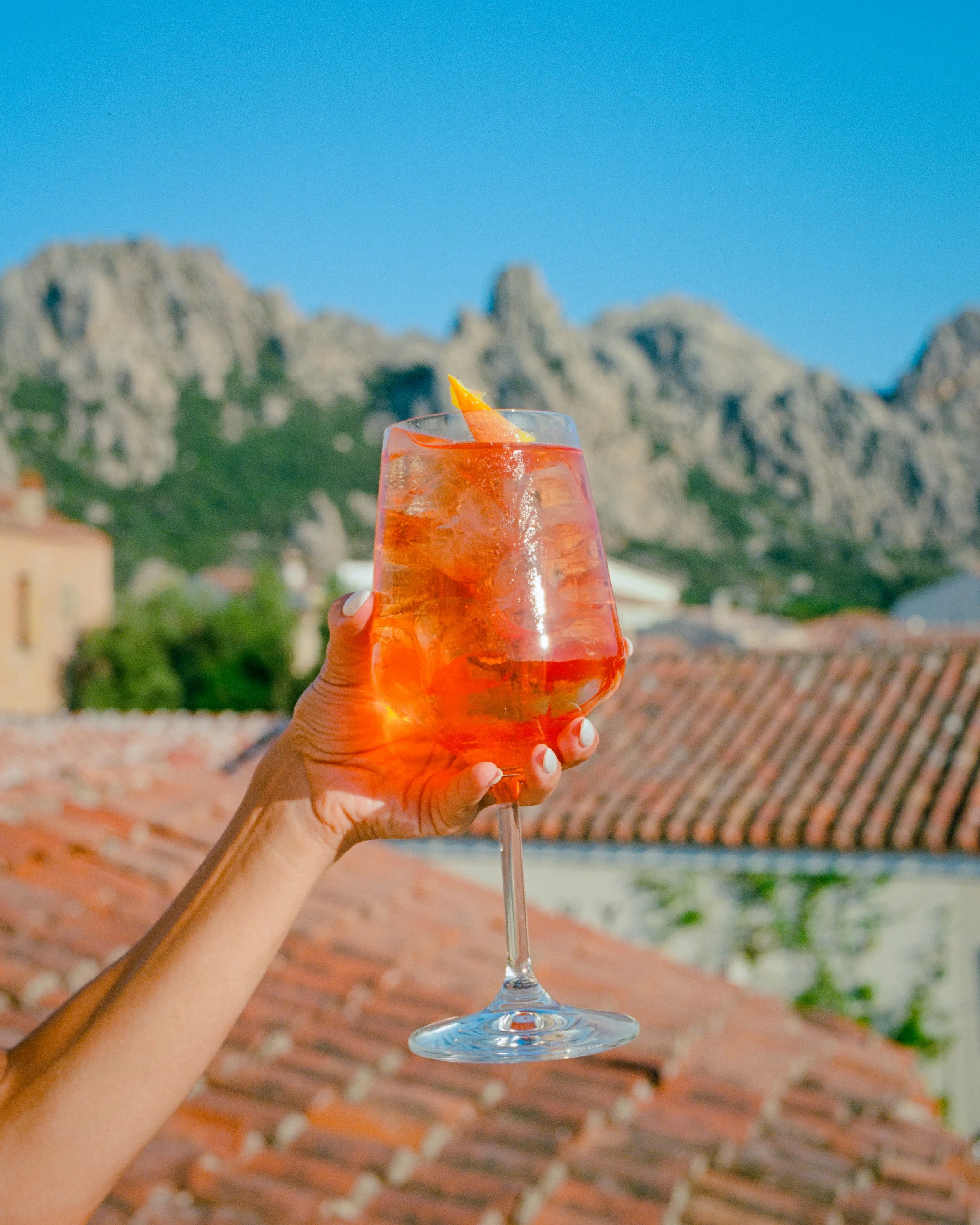 A hand holds up a spritz above terracotta roofs, granite peaks behind