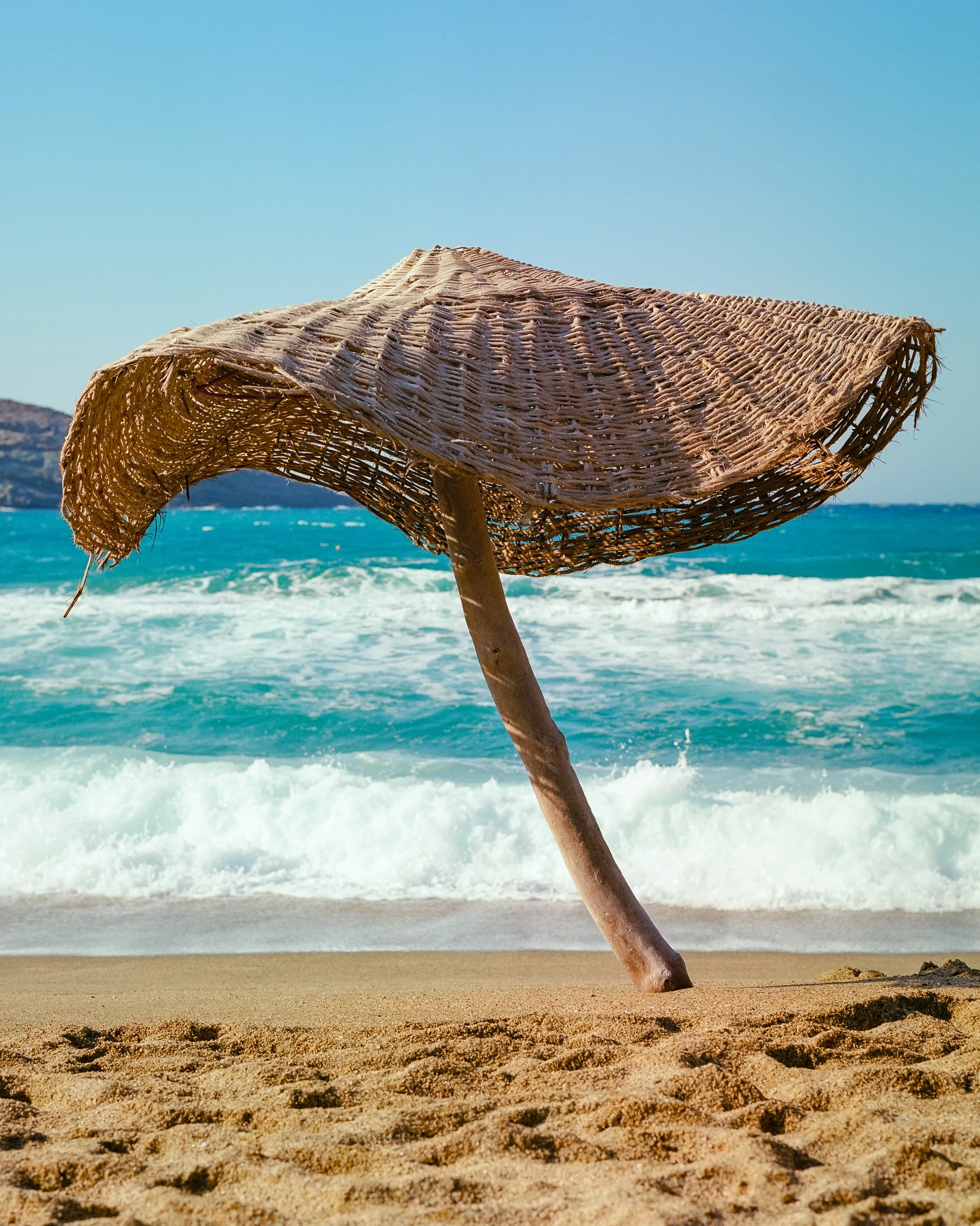 A woven straw beach umbrella leaning on a driftwood pole, with turquoise surf breaking behind it