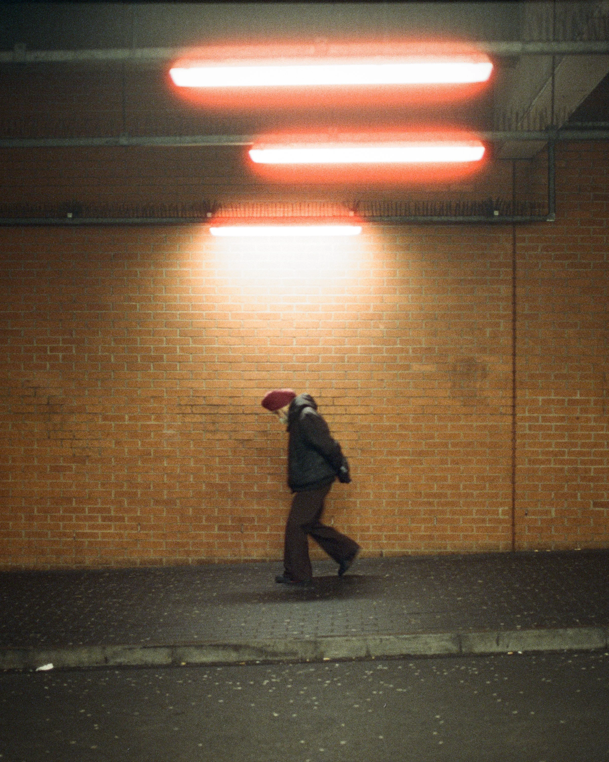 A man walking under three glowing strip lights on a brick wall