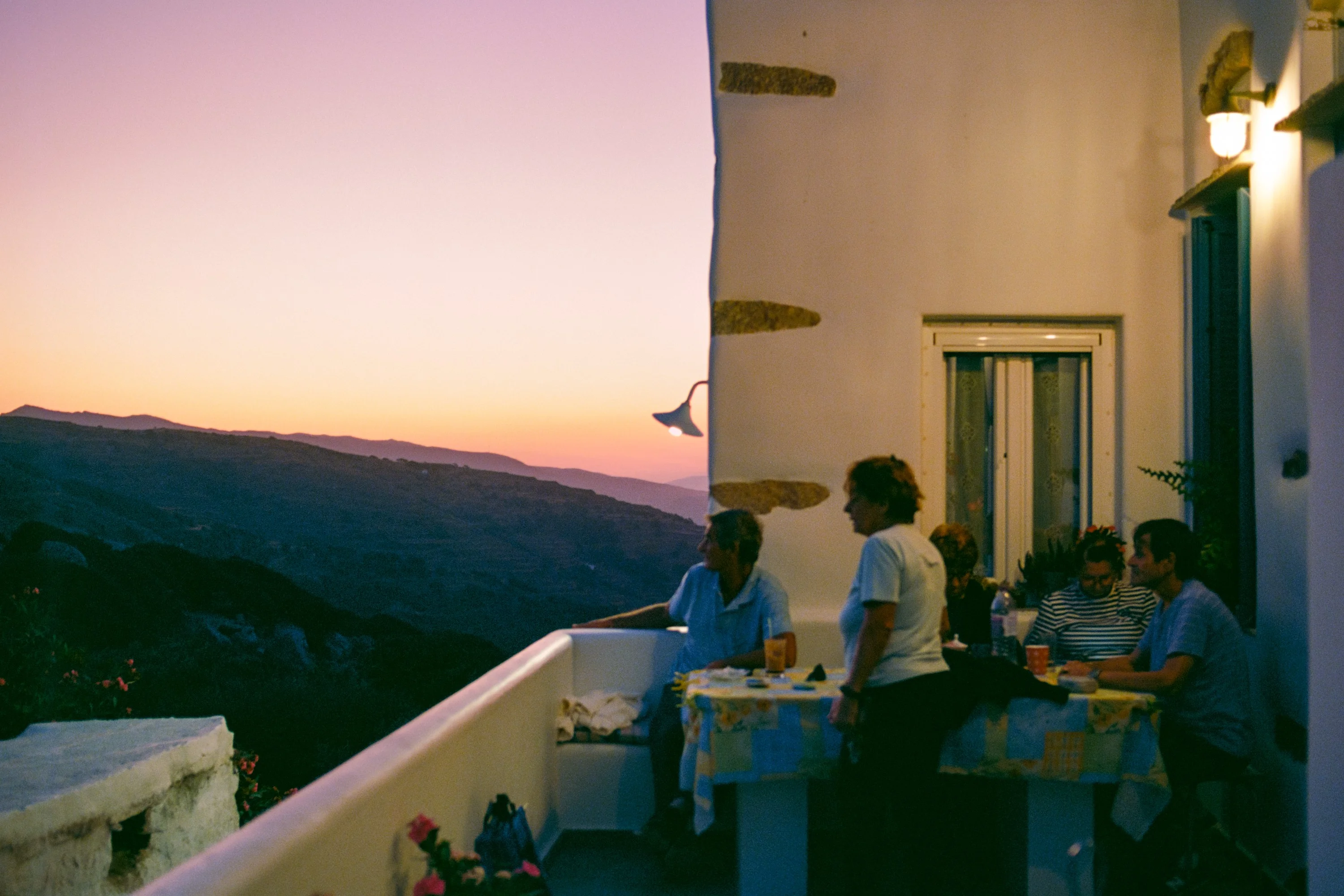 People at a taverna table on a terrace at dusk, mountains fading behind
